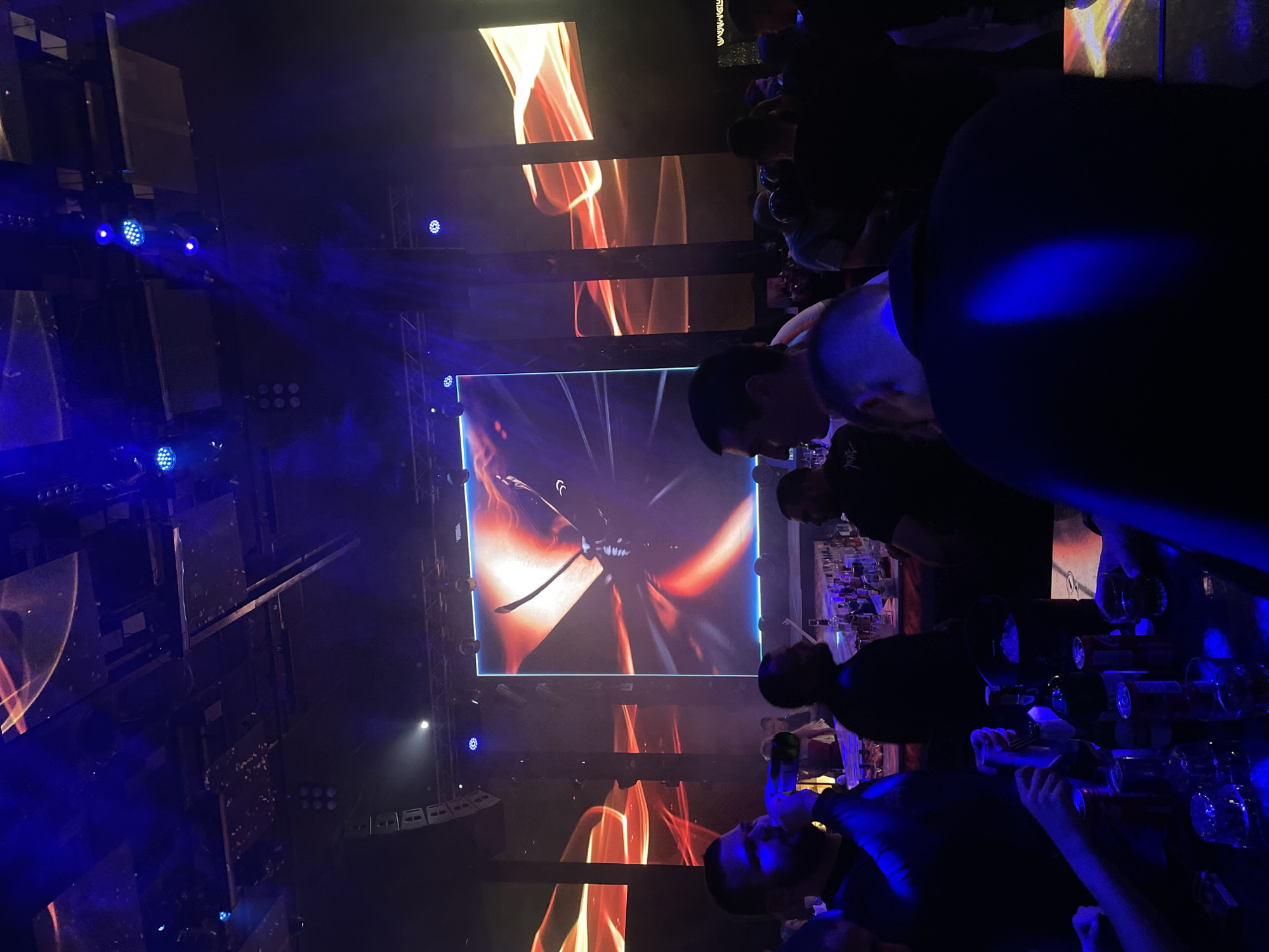 This image captures a lively scene inside an indoor venue, likely a club or event space, at night. The atmosphere is dark and energized, dominated by vibrant artificial lighting and large screens.
In the center, a large, rectangular LED screen displays a striking visual of what appears to be a stylized sword or propeller against a fiery, abstract background with shades of orange and red. This central display is flanked by multiple smaller LED panels on the surrounding walls and ceiling, which also show similar abstract, fiery patterns, creating a cohesive visual theme. Numerous blue LED spotlights are strategically placed throughout the venue, casting beams of light and adding to the dynamic ambiance. Some white spotlights are also visible. Sound equipment, possibly large speakers, are mounted on structural frames on the left side of the image.
Several people are visible, mostly men, gathered in the space. In the immediate foreground, a man is looking upwards, holding a green bottle, suggesting he is enjoying the event. Further back, other individuals are standing, observing the screens or interacting. To the right, a bar-like area is visible, with shelves holding bottles and glasses, indicating a social gathering or party environment. The entire scene is bathed in a mix of blue and red light emanating from the various displays and light fixtures. No discernible text is visible on the screens or elsewhere. The overall impression is one of an exciting nightlife event taking place in Plovdiv, Bulgaria.