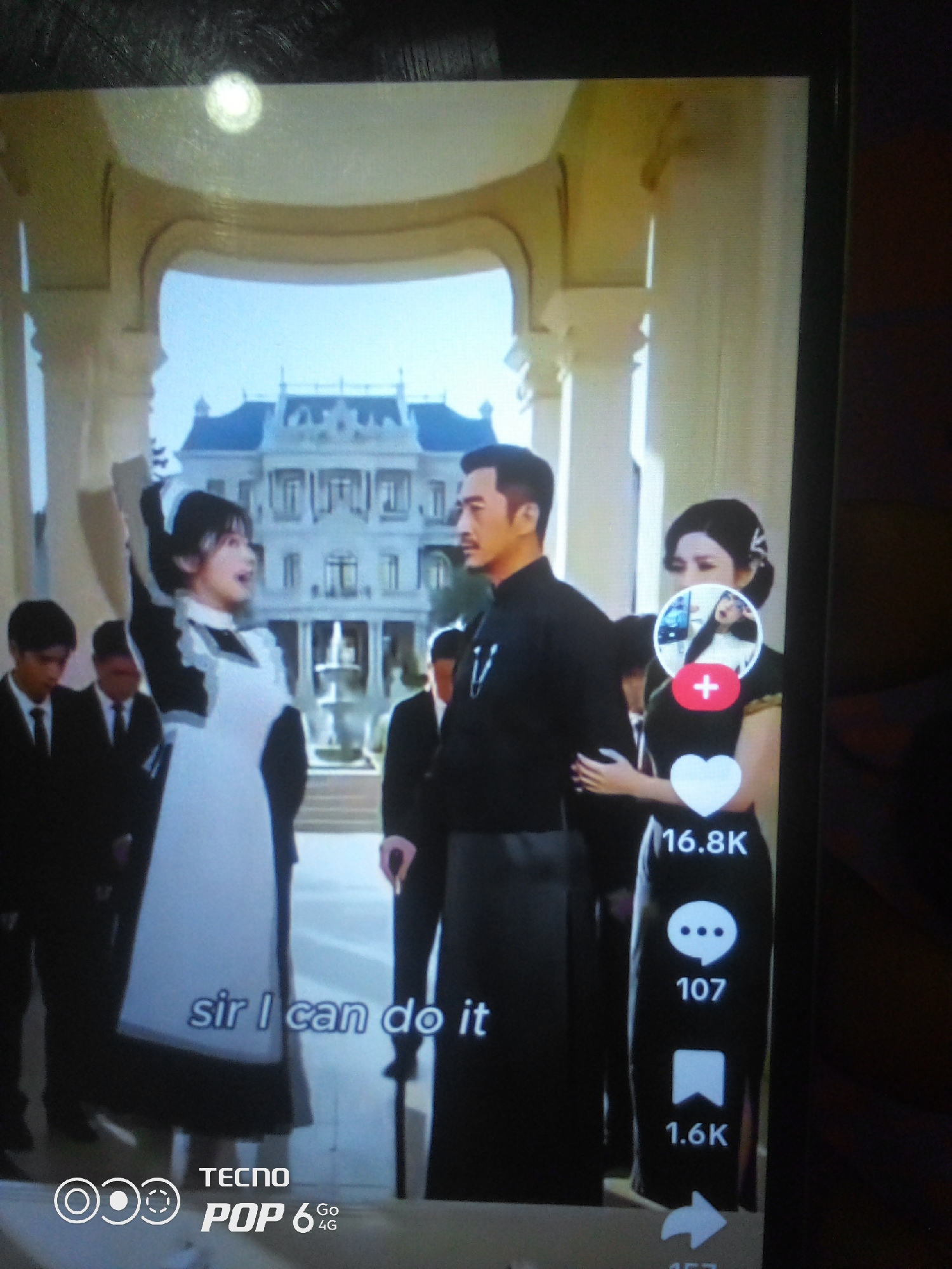 In an outdoor setting, likely a grand estate, with a large, opulent building in the background, a woman in a maid's uniform stands to the left of a man in a traditional Chinese garment. She is holding a white cloth or paper above her head, gesturing upwards. Another woman in a cheongsam, with a small circular image superimposed on her chest displaying a person taking a selfie, stands to the right of the man. Below the woman in the cheongsam are social media icons and engagement metrics: a heart with "16.8K" below it, a speech bubble with "107" below it, and a bookmark icon with "1.6K" below it. In the lower left corner, the text "sir i can do it" is visible. The TECNO POP 6 4G logo is also present at the bottom of the image.