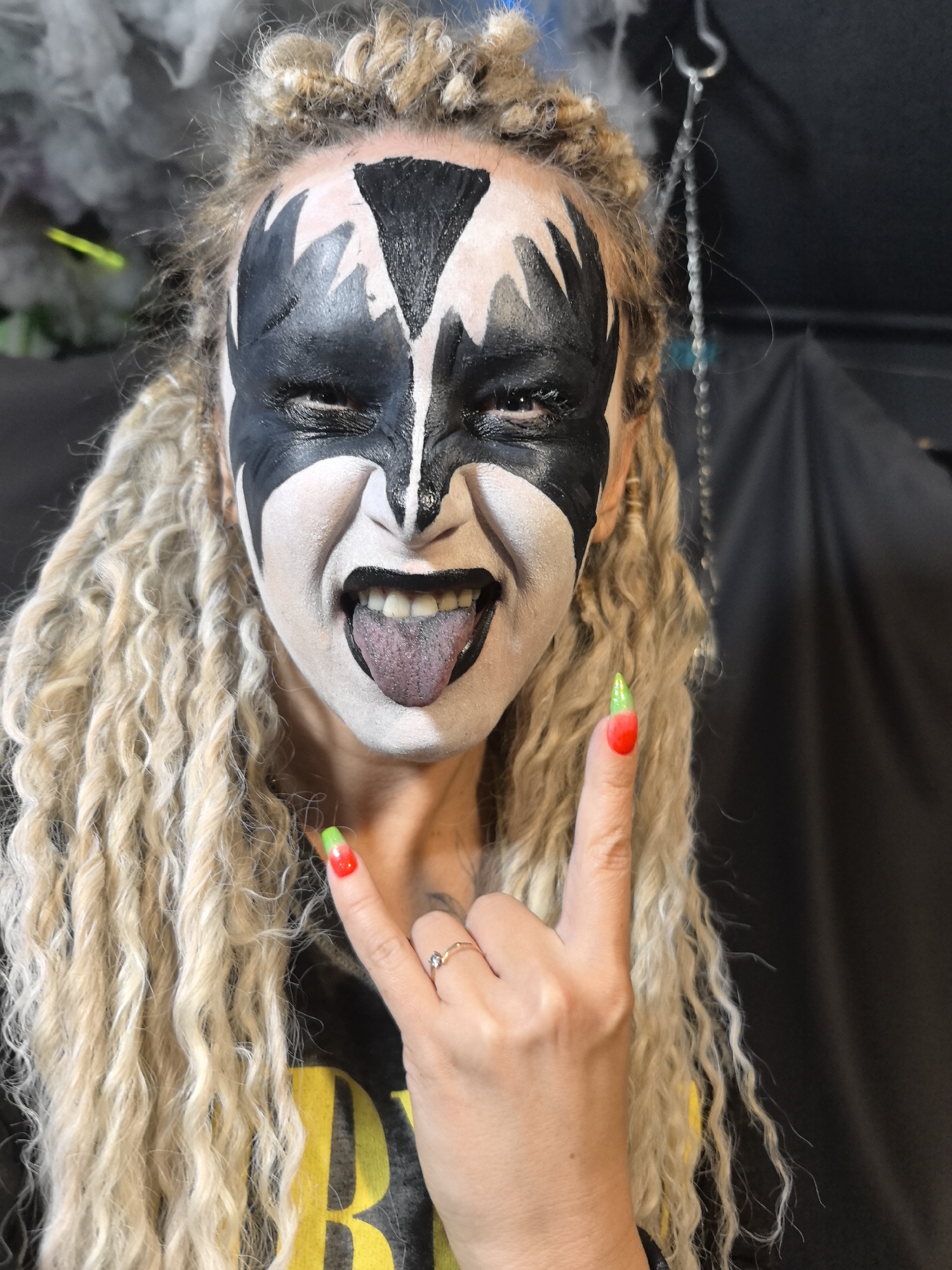 The image captures a person adorned with detailed black and white face paint, styled to resemble the "Demon" character from the band KISS. The subject has long, light blonde, tightly curled or dreadlocked hair that cascades around their face and shoulders. They are shown in an energetic pose, with eyes squinted, mouth open wide, and tongue sticking out.
The person's right hand is raised, making the "devil horns" gesture, a symbol commonly associated with rock and roll music. Their fingernails are long, pointed, and painted in a striking red with bright green tips. A small ring with a clear stone is visible on the ring finger. The subject is wearing a dark-colored shirt, with a partial glimpse of yellow text or a design visible on the left side of the chest, which is not legible.
The background is dark and indistinct, suggesting an indoor or shaded outdoor setting. A blurry light-colored element is visible on the upper left, and a thin, chained object hangs from the top right of the frame. The overall impression is one of a vibrant, themed event, concert, or party. No specific location cues for Noviy, Russia, are discernible from the image itself, nor is the time of day explicitly indicated. The prevailing emotion conveyed is one of playful ferocity and rock-and-roll enthusiasm.