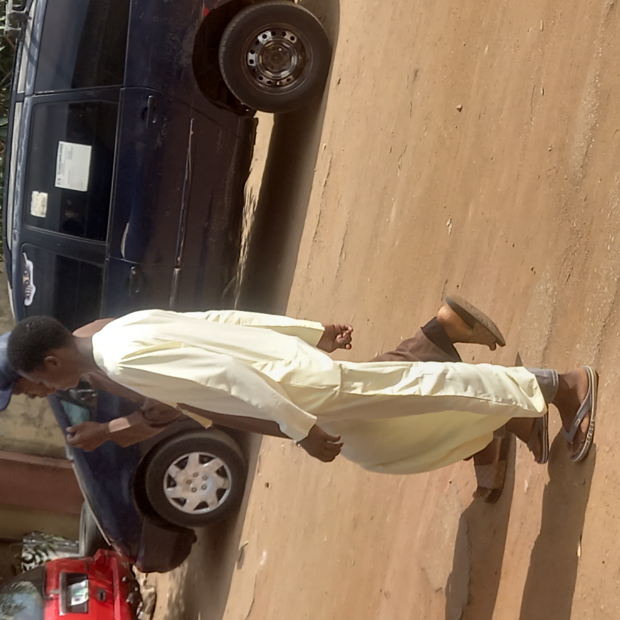 The image shows two men walking on a dirt road in Dadi Riba, Nigeria. A dark-colored van is parked partially on the road, near the top of the image. A red car is visible at the bottom left. One man is wearing a light-colored, full-length garment; the other is in brown clothing. Both are wearing sandals. The road is dirt and dusty.