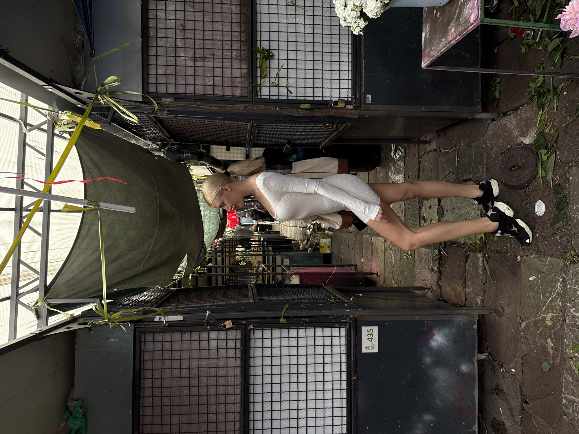 The image depicts a woman in a white dress and black sneakers walking down an alleyway. The alleyway appears to be an outdoor market in Vracar Urban Municipality, Serbia, given the stalls on either side, the cobblestone ground, and the foliage visible. Some stalls are enclosed with metal mesh, and various plants are visible, including cut flowers. The scene is somewhat cluttered, but the woman is the central subject.