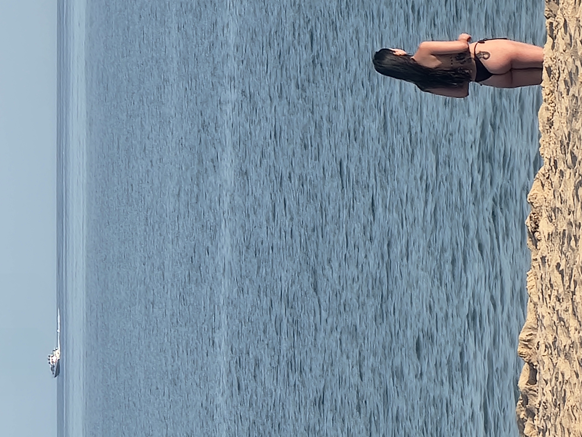 A woman stands on a sandy beach, facing away from the camera and looking out at the ocean. She is wearing a black bikini. In the distance, a white boat with several people on board is moving across the water. The sky is clear and the ocean is calm. The scene appears to be located in Quarteira, Portugal, a popular tourist destination known for its beaches and seaside promenade. The foreground is dominated by the sandy texture of the beach, with gentle undulations. The midground is the expanse of the ocean, with subtle ripples on its surface. The background features the horizon line where the ocean meets the pale blue sky, and a small boat.