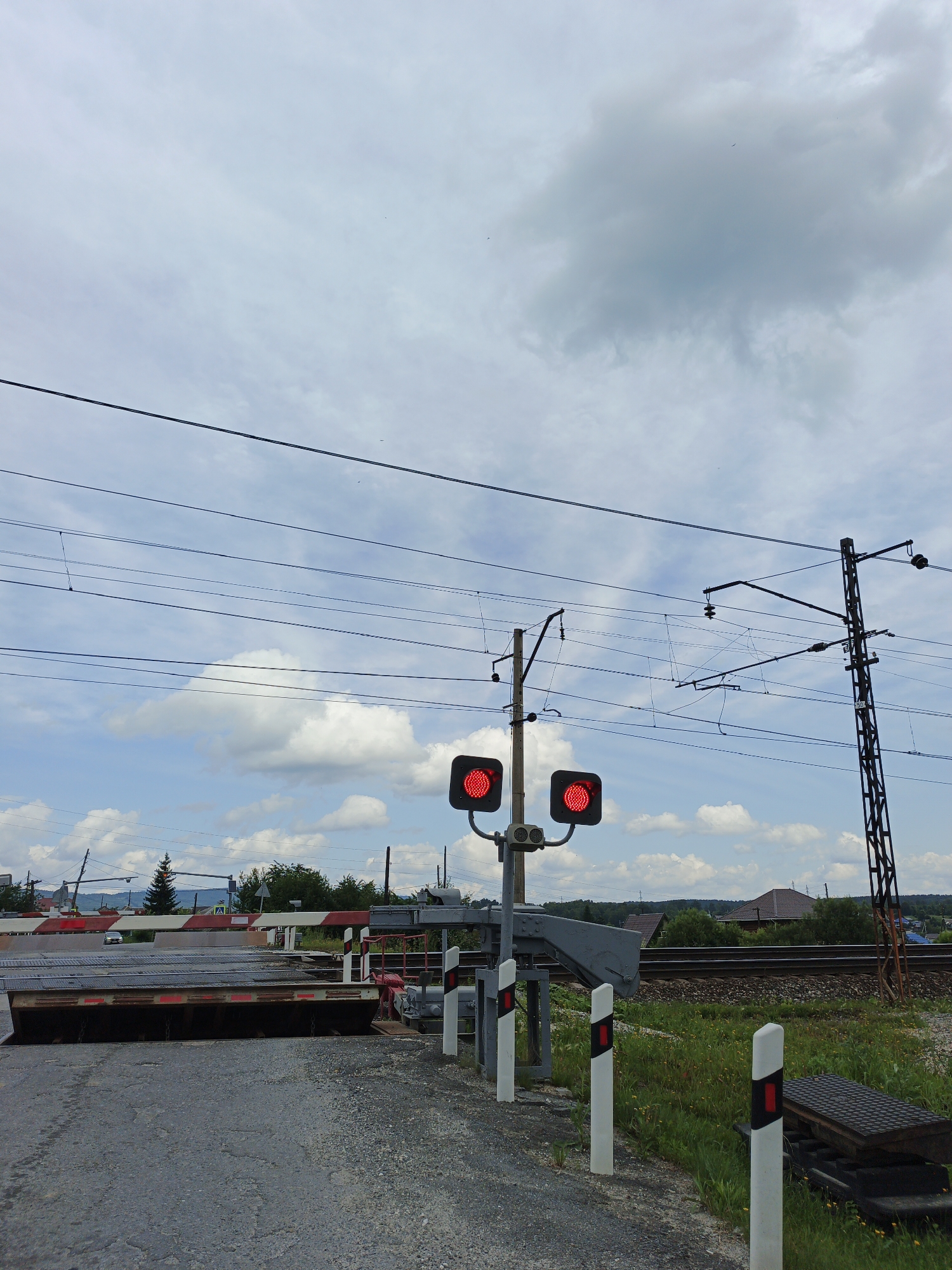 The image captures a railroad crossing in Medvedyovka, Russia, on a cloudy day. A closed railroad crossing barrier, painted with red and white stripes, is prominently displayed in the foreground. Two traffic lights, both displaying red signals, are mounted on poles. Overhead, a complex network of electrical wires, likely for the railway, crisscrosses the sky, supported by utility poles. To the right, a tall, dark metal tower stands, also part of the electrical infrastructure. Beyond the crossing, railroad tracks are visible, and the background features scattered houses, trees, and a gentle slope of land. The ground is paved with asphalt and gravel. There are no people visible in the frame.