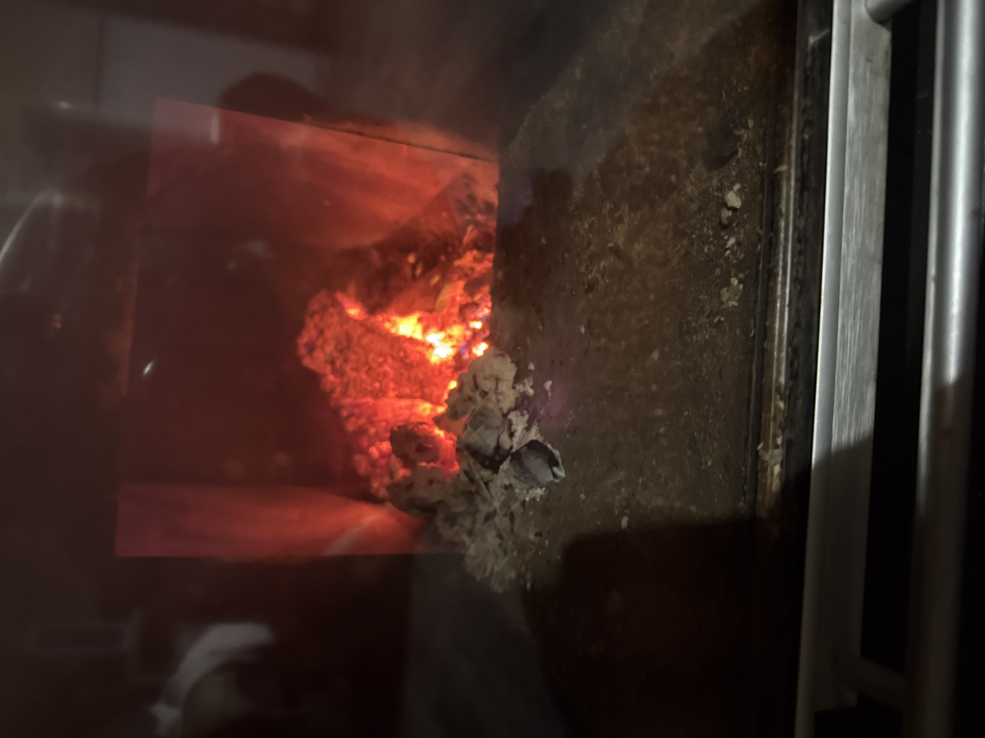 The image shows a wood-burning stove in operation. The interior of the stove contains burning wood with visible flames and glowing embers. There is a dark, textured surface adjacent to the firebox, likely the interior wall of the stove. The stove door, or a portion of it, is visible, with metal bars or framing on the right side of the image. The view is from outside the stove, looking in through the glass window, which is slightly reflective. The location is not discernible from the image.