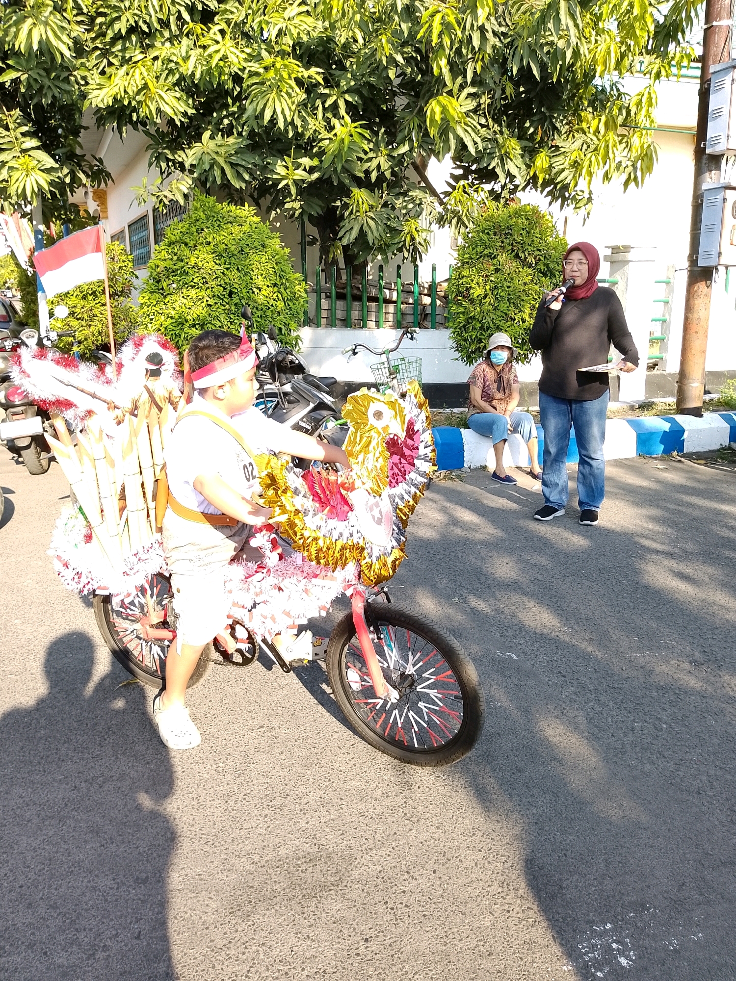 The image depicts an outdoor scene in Surabaya, Indonesia, likely during a public event or parade. In the foreground, a young boy is positioned next to a bicycle decorated with colorful streamers, bamboo stalks, and what appears to be a festive headdress. He is wearing a white t-shirt, shorts, and white shoes. The bicycle itself is adorned with red and white accents on its spokes.
Behind the boy, a woman wearing a headscarf and a black sweater, holding a microphone, is speaking. She is standing on the asphalt road. To her left, another person, possibly a woman, is seated on a blue and white curb, wearing a patterned shirt, jeans, and a face mask.
The background features lush green trees and a building with white walls. The presence of a red and white Indonesian flag suggests a patriotic celebration. Several motorcycles are parked in the background, indicating a public gathering. The lighting suggests it is daytime, with shadows cast on the road, indicating sunlight.