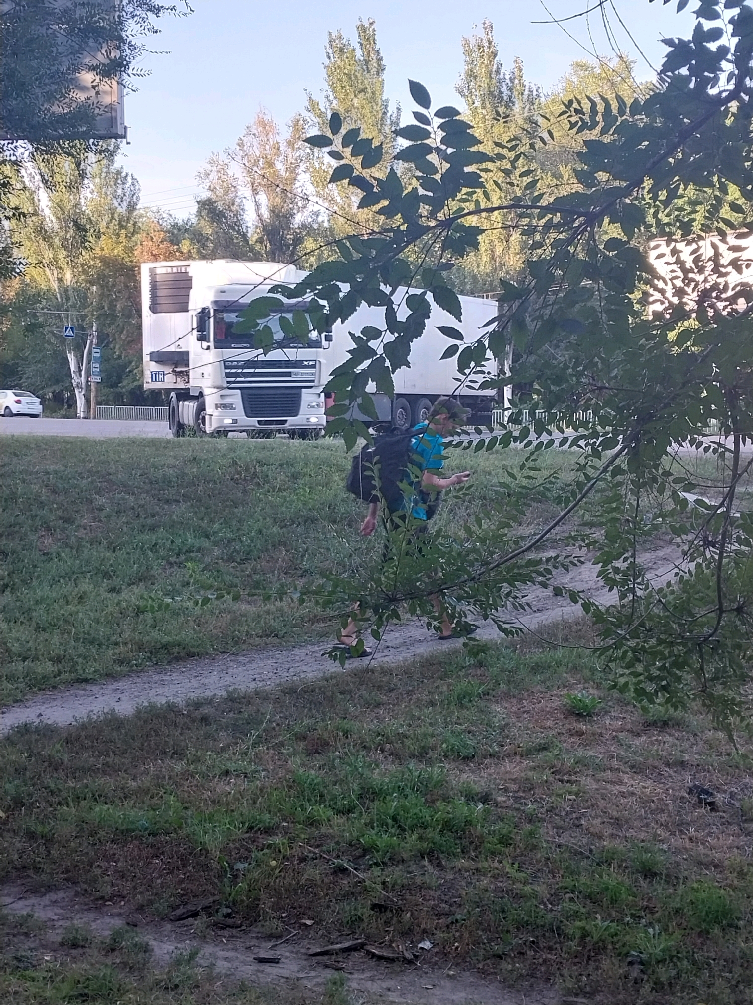 The image shows a young man with a black backpack walking on a dirt path next to a grassy area. He is wearing a blue t-shirt and dark shorts, and appears to be holding something in his left hand. In the background, a white semi-trailer truck is parked on a road. Trees and foliage are visible throughout the scene. The location appears to be a road in Dnipro, Ukraine, near the Dnipro River. The lighting suggests it is daytime, possibly late afternoon due to the shadows.