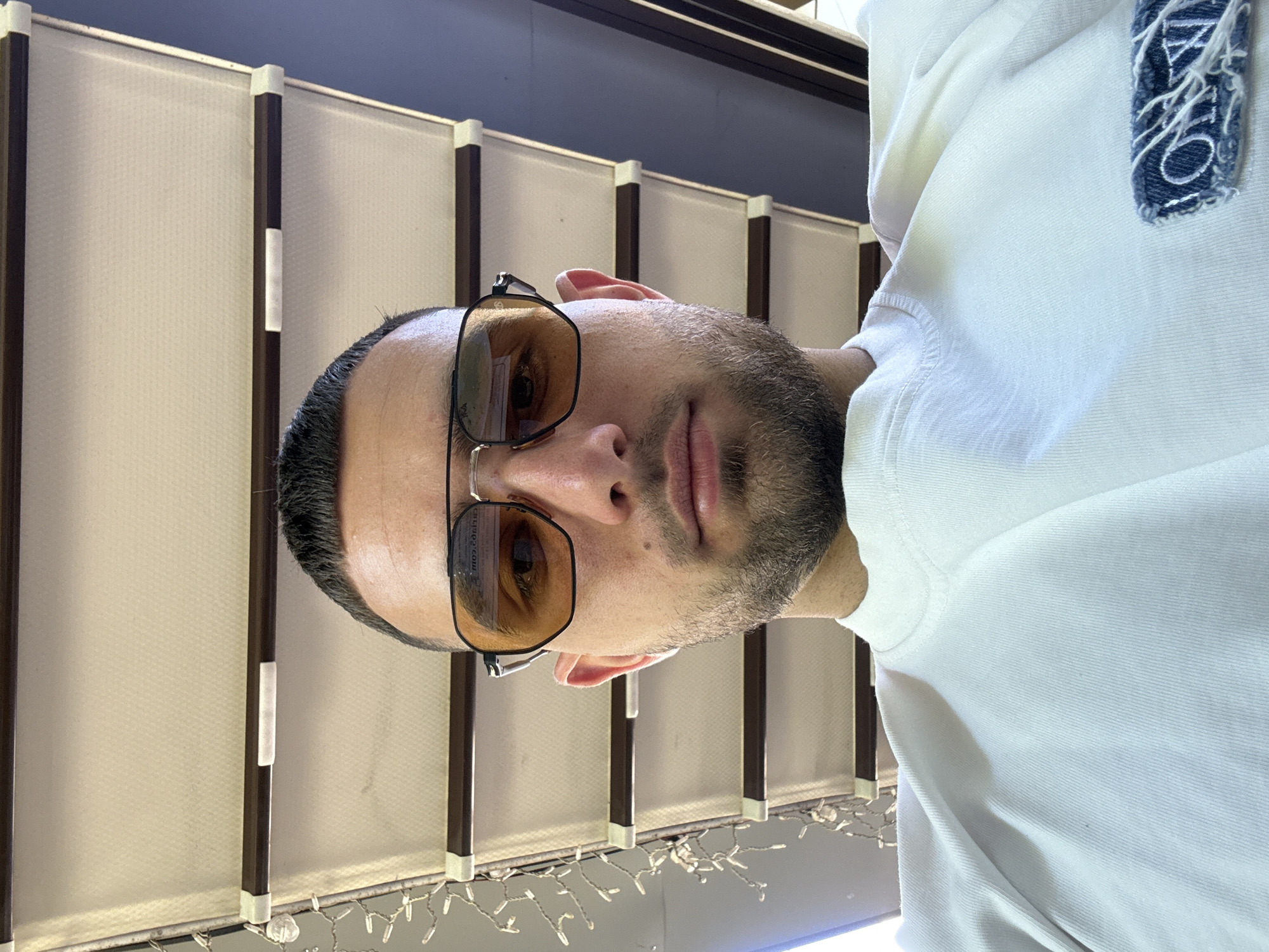 A man with short dark hair, a trimmed beard, and wearing gold-tinted eyeglasses is positioned facing the camera. He is wearing a white, possibly cotton, long-sleeved shirt or sweater. The background consists of vertically aligned beige panels with dark brown frames. A string of lights is partially visible beneath the panels. The location appears to be indoors near a wall or partition. There is no explicit information within the image that directly confirms it is in Stara Zagora, Bulgaria.
