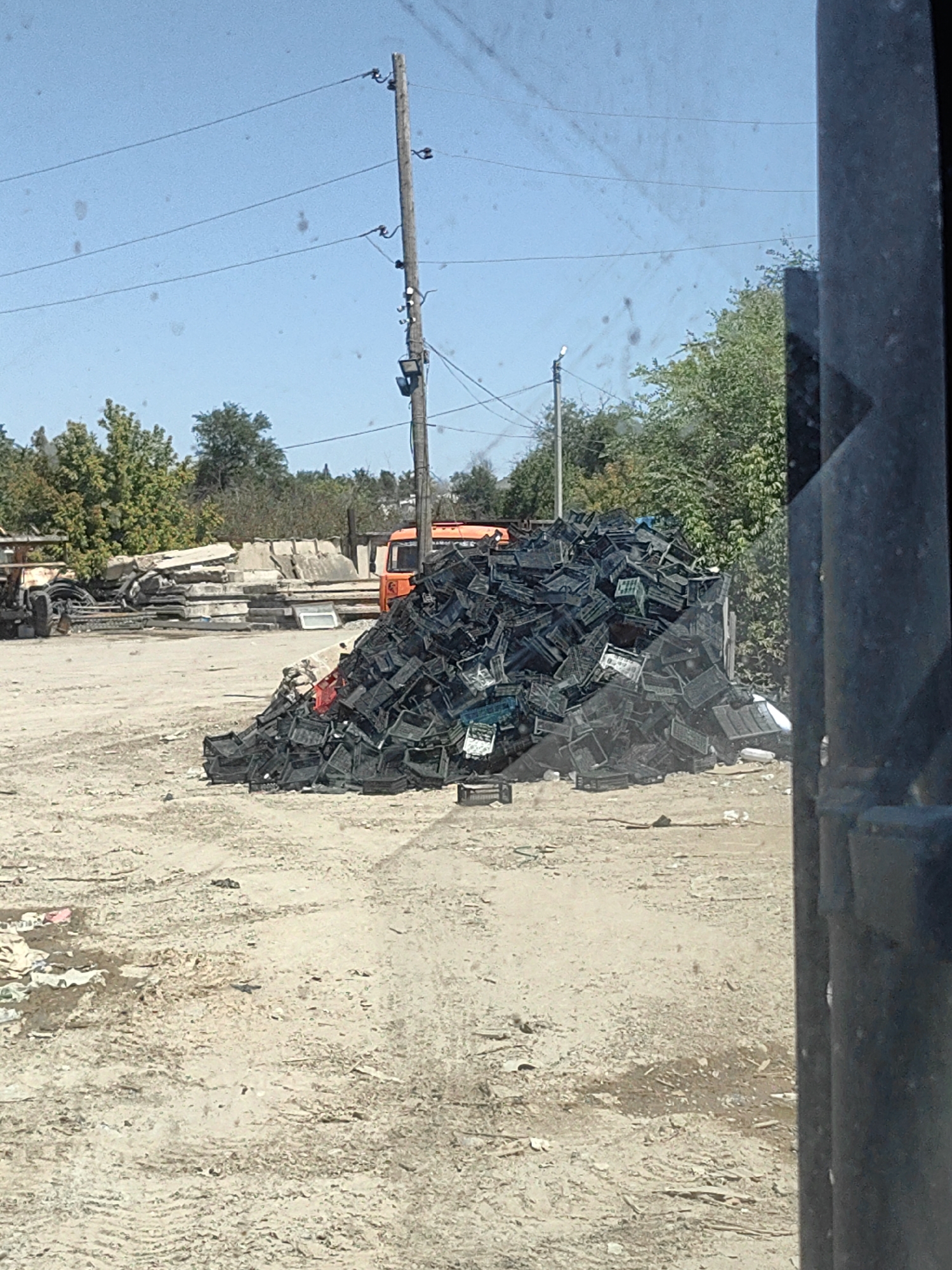 The image shows an outdoor scene with a large pile of black plastic crates in the foreground. Behind the crates, there is an orange truck, piles of concrete slabs, and trees. In the background, there is a wooden utility pole with power lines. The ground is covered in dirt and debris. The scene appears to be a junkyard or recycling facility. The location is the city of Shymkent, Kazakhstan.
