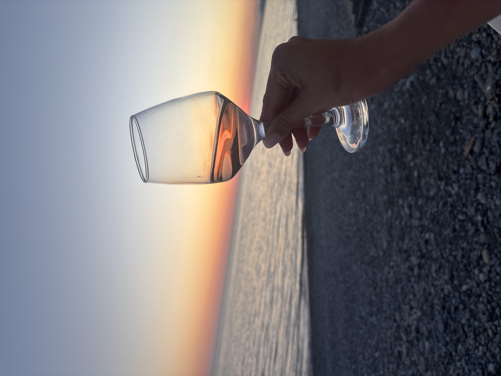 A human hand holds a clear wine glass containing a light-colored liquid, likely rosé, against a sunset over the Black Sea in Sochi, Russia. The sky transitions from a vibrant orange-red at the horizon line, where the sun is setting, to a pale blue-grey at the top of the frame. The liquid within the glass and the glass itself reflect these warm hues, showing distinct orange and pink tones. Small condensation droplets are visible on the outer surface of the glass. The hand, with neatly manicured nails, grasps the stem of the glass, which is positioned in the mid-right portion of the image. The calm sea stretches across the mid-ground, reflecting the subdued light of dusk. The immediate foreground consists of a dark, out-of-focus pebble beach.