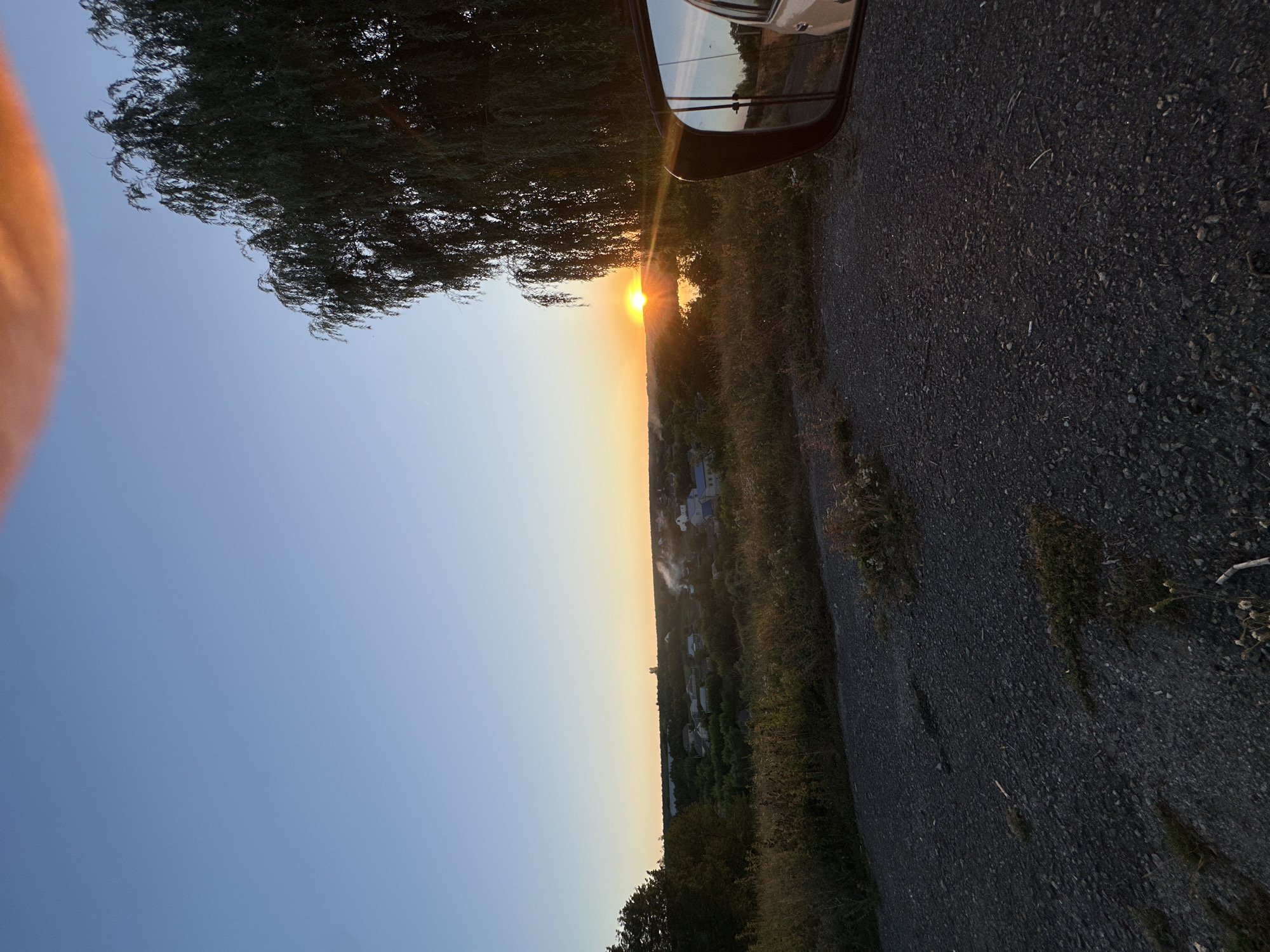 The image depicts a sunset over a rural landscape, likely the outskirts of Makiv, Ukraine. A gravel road occupies the foreground, with dry grass and weeds growing along its edges. In the midground, a village with houses and trees is visible, with smoke rising from a few chimneys. A body of water, possibly a river or lake, is situated behind the village, reflecting the warm hues of the sunset. A large weeping willow tree stands on the right side of the frame, its branches casting shadows. A car's side mirror is also visible on the right, reflecting a portion of the scene, including the road and the willow tree. The sky is clear and blue, with a soft orange glow near the horizon where the sun is setting.