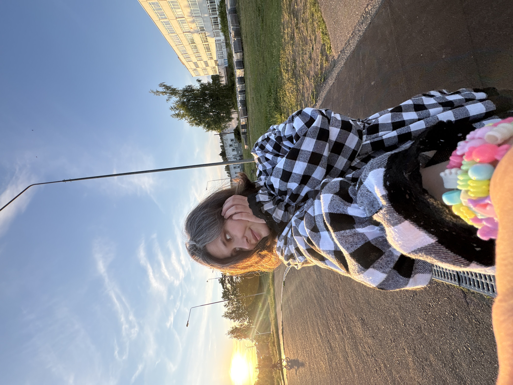 This media file is a selfie of a young person with grey-streaked hair, wearing a black and white checkered shirt. They are looking at the camera, with one hand resting on their cheek. The person is sitting outdoors, with a sunset visible in the background. The sky is a clear blue with wispy clouds. In the distance, there is a large, multi-story building and trees. The ground is paved with asphalt and there is a metal grate in the foreground. A colorful beaded bracelet is visible on the person's wrist. The time of day is late afternoon or early evening, as indicated by the setting sun and warm light. The weather appears to be clear and mild. The location is likely Chaikovsky, Russia, given the context. There is no visible text in the image.
