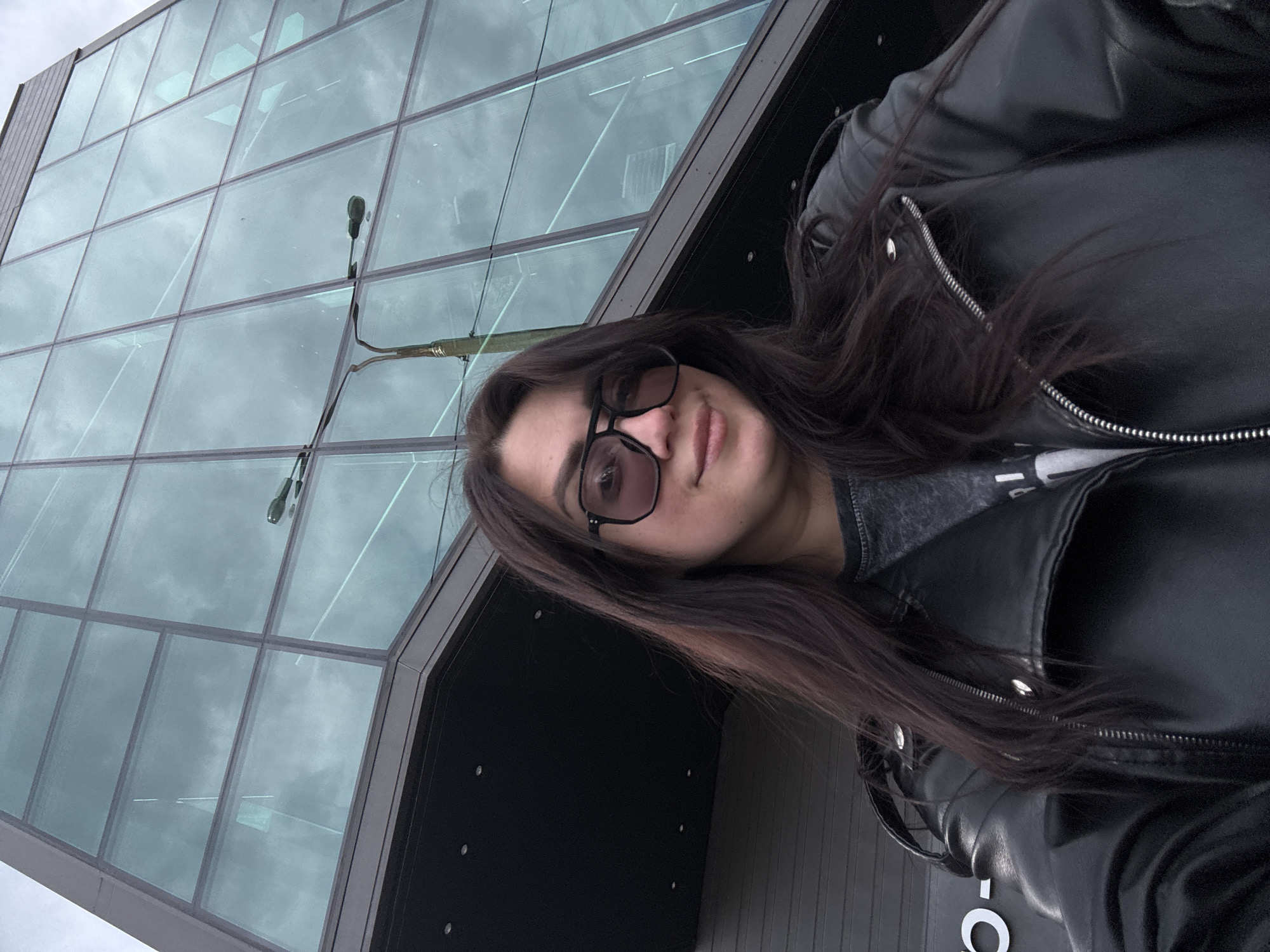 A medium shot shows a female wearing sunglasses and a black leather jacket, positioned in front of a modern building with a glass facade. The sky reflects in the glass panes. A street light stands adjacent to the building. The location is in the city of Unknown, Unknown.