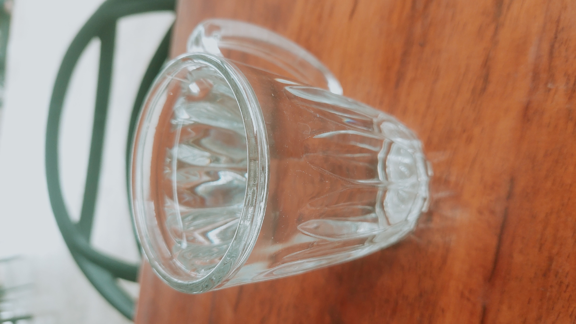 A clear glass cup filled with water sits on a wooden table. The cup has a ridged pattern and a small handle. In the background, a blurred green object, possibly part of a chair, is visible against a light-colored wall. The image was taken in Meunasahmee, Indonesia.