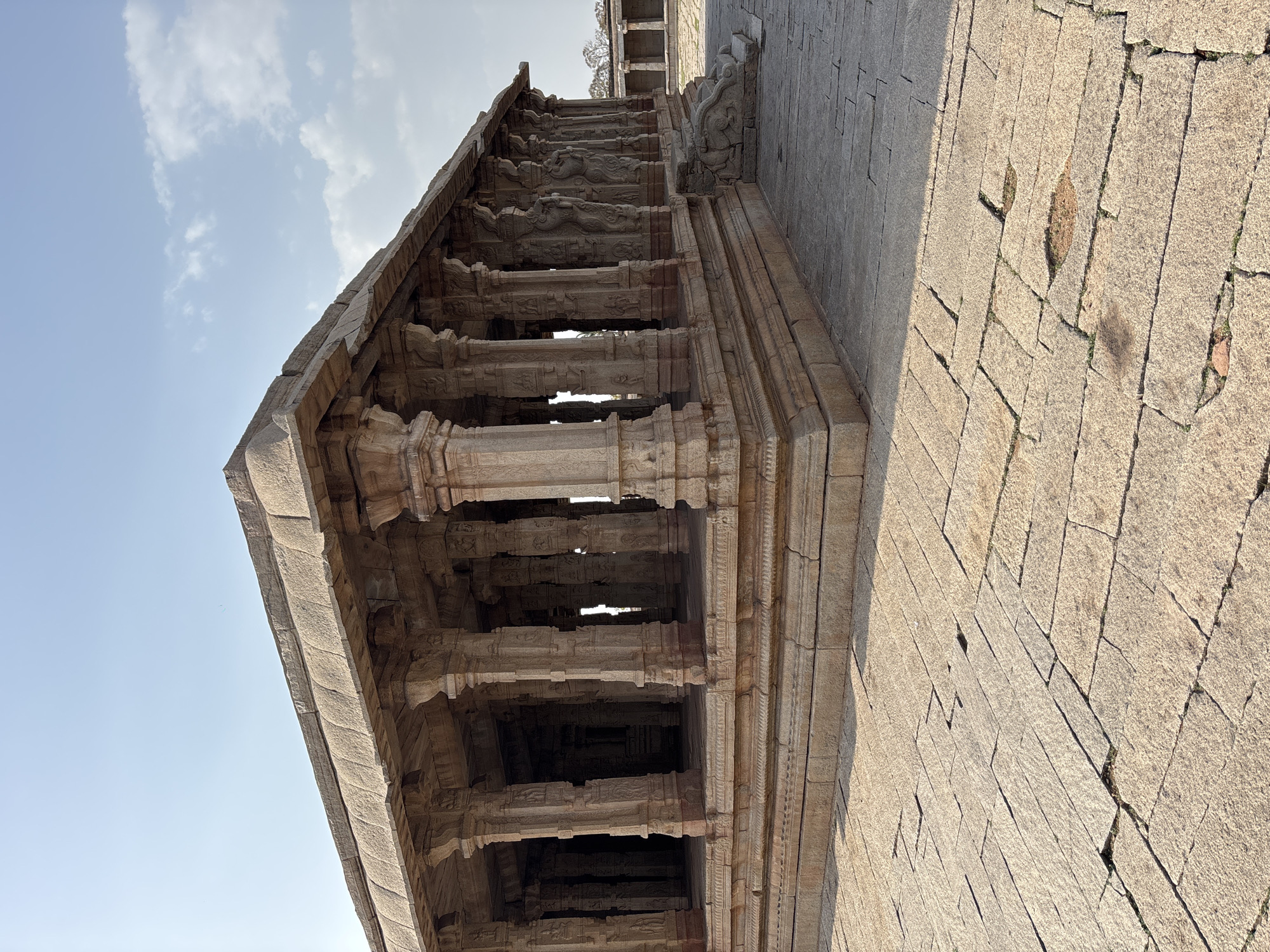 The image captures a low-angle, side-view of a stone structure located in Powerhouse Colony, India. The structure appears to be an ancient temple or building, exhibiting intricate stone carvings and stacked layers. The columns and walls are made of aged, textured stone. The foreground shows a stone-tiled pathway with cracks and irregular patterns. The sky, visible in the upper-left, is light blue with scattered white clouds. The overall scene conveys a sense of historical architecture and weathered stonework.