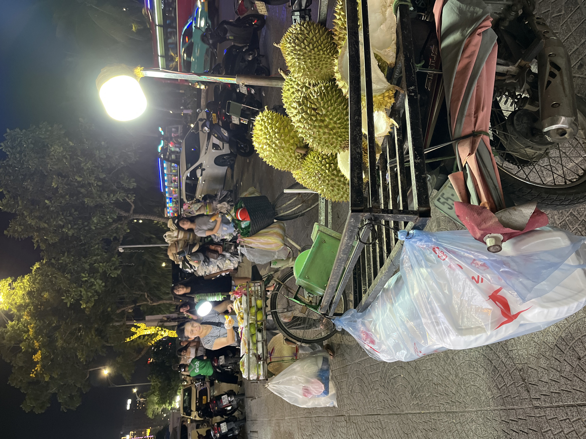 The image shows a street scene at night in Đà Nẵng, Vietnam. A durian vendor is operating from a bicycle stand on the sidewalk, with several ripe durians displayed on the stand. A woman, likely the vendor, is attending to customers. An array of motorcycles and scooters are parked along the curb, typical of the traffic in Vietnam. The surrounding area is lit by streetlights and decorative lights, creating a lively atmosphere. People are walking and congregating nearby. The scene shows everyday street commerce.