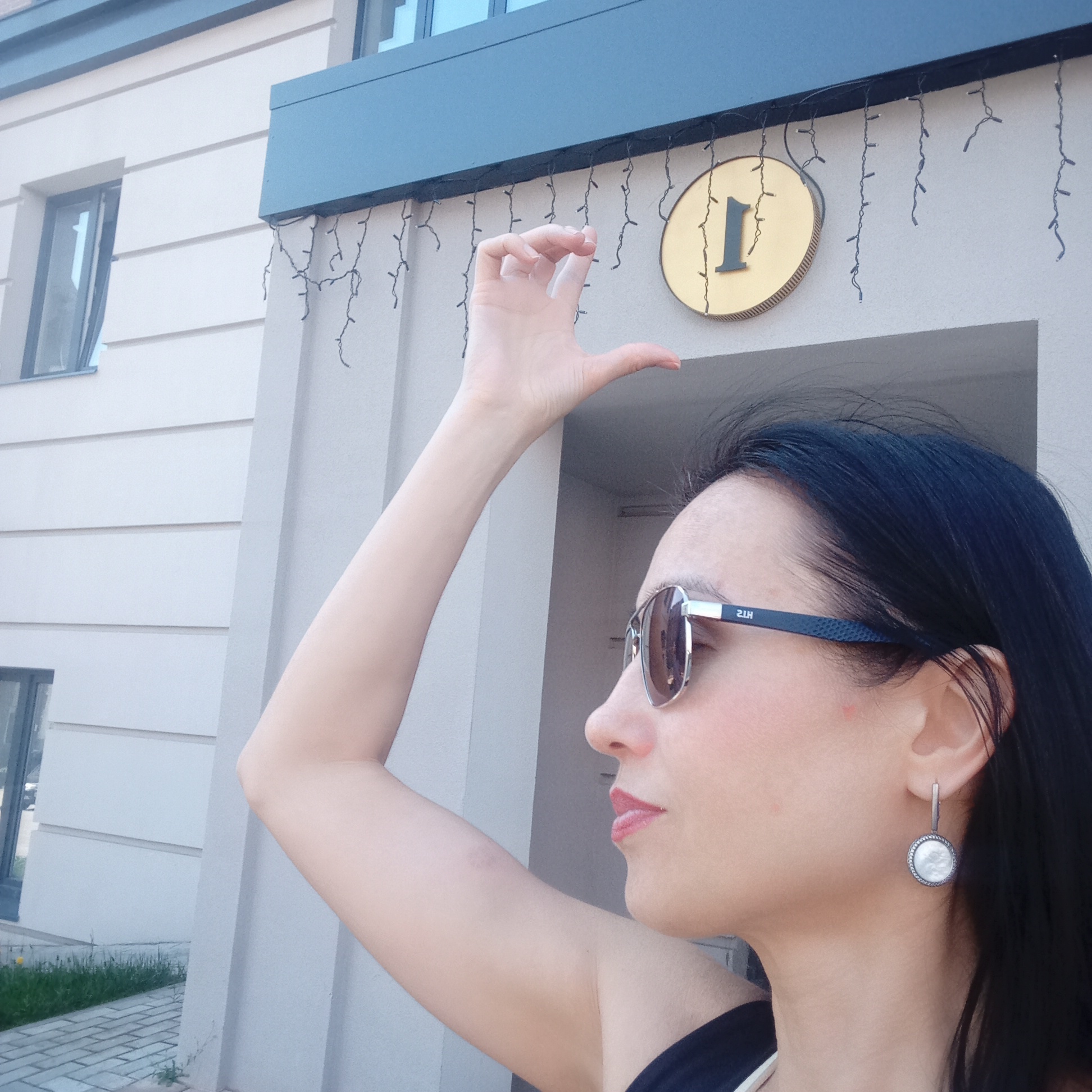 A woman is standing in front of a building at address number 1, possibly in Pervomaysky, Russia. The woman is in profile, facing left. She is wearing sunglasses and an earring. Her arm is raised, and her hand is positioned near the address number sign. The building is painted in light colors, with decorative lighting attached above the number.