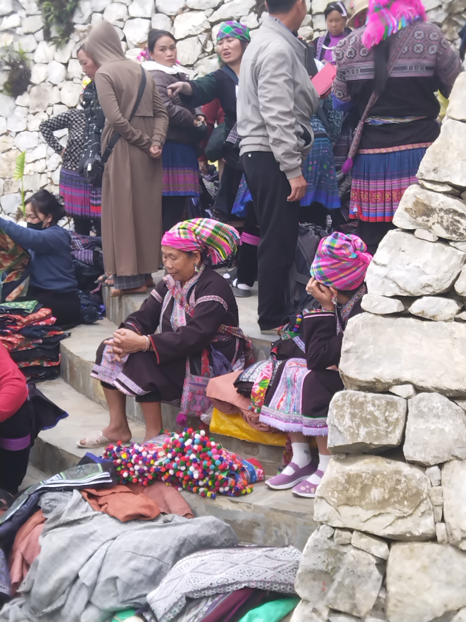 In an open-air setting, possibly a marketplace in Unknown, Unknown, a gathering of people is seen. The background includes what appears to be a stone wall and steps. Several individuals are clad in traditional attire with vibrant colors and intricate patterns, including headwraps. An older woman with a colorful headwrap is seated on the steps, and a younger person in similar attire sits beside her. Merchandise, possibly textiles or handicrafts, is displayed on the steps and in front of the seated women. Other people in the background are wearing modern clothing, suggesting a mix of locals and visitors. Interactions between people suggest a commercial transaction or social gathering.