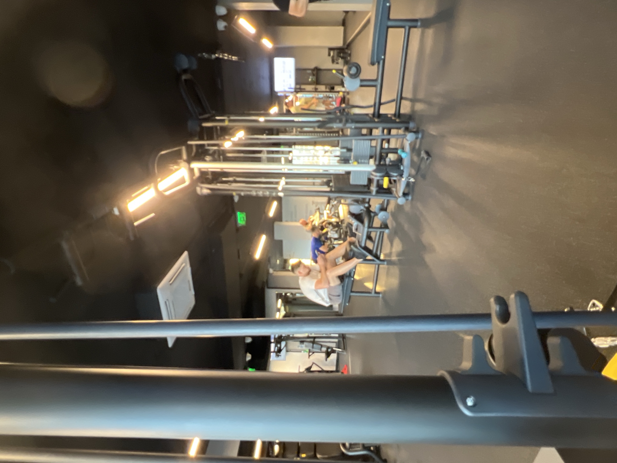 The image, which is rotated 90 degrees clockwise, depicts a modern indoor gym. In the foreground, large, dark metal bars from a piece of equipment are out of focus. The main subject is a woman, wearing a white top and light-colored shorts or leggings, seated on a leg press machine in the mid-ground, appearing to be exercising or resting. Further back, another individual in a blue top is partially visible, also engaged with gym equipment.
The gym space is equipped with numerous cable machines arranged in rows. The ceiling is dark and features several bright, rectangular strip lights and circular spotlights that emit a warm, yellow light, casting prominent shadows on the dark floor. A white air vent is also visible on the ceiling. In the distant left, a small green illuminated "EXIT" sign can be seen. The overall scene suggests a typical fitness center during operational hours. There is no legible text other than the "EXIT" sign.