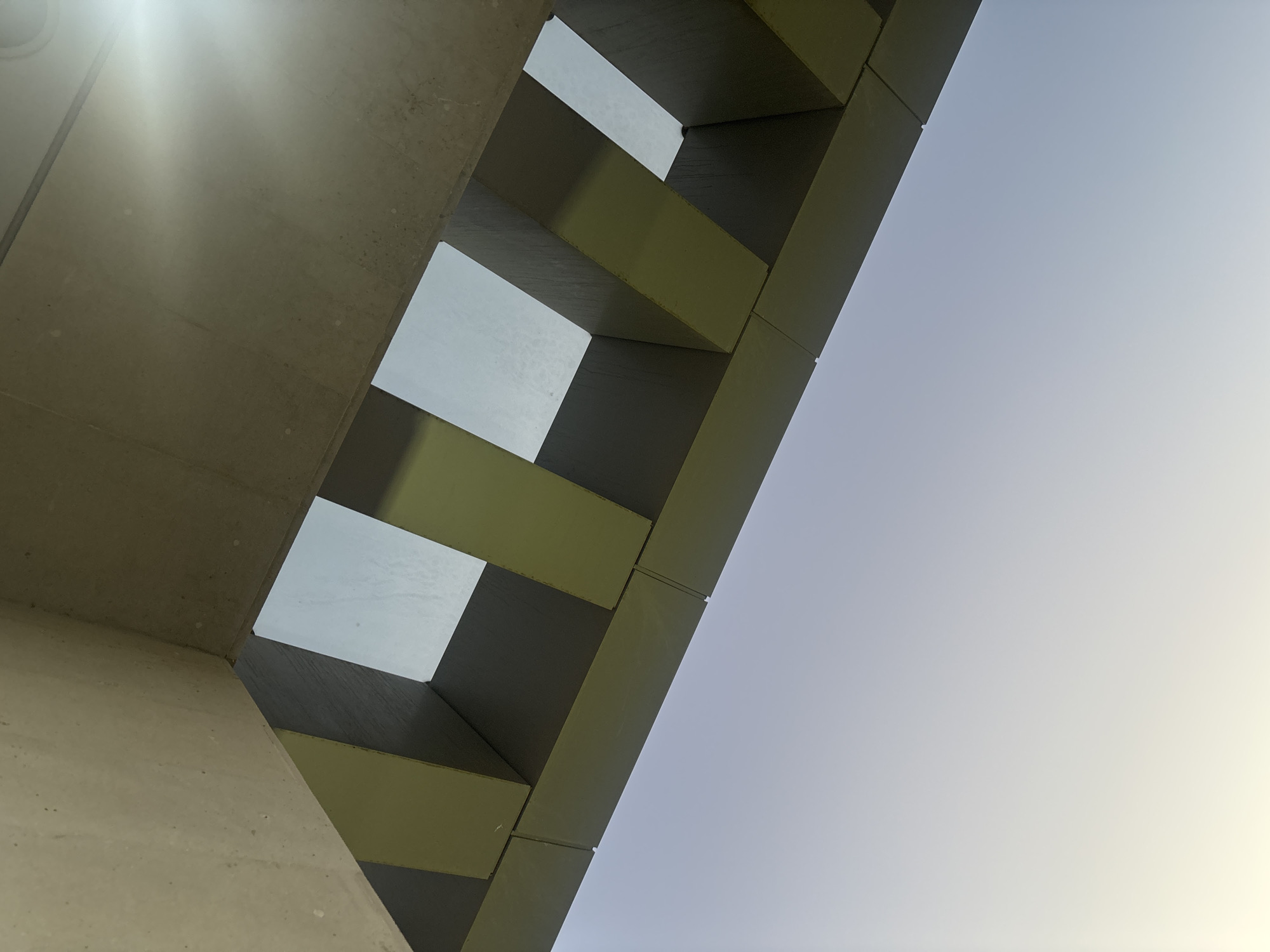 The image presents a low-angle, abstract view of a modern architectural structure, looking upwards towards the sky. The scene is dominated by a clear, bright daytime sky, which transitions from a light blue in the upper left to a pale yellow or white hue towards the right.
In the foreground and extending from the bottom left, a rough-textured, light-beige concrete surface is visible, brightly illuminated by direct sunlight, causing a distinct lens flare in the upper left quadrant. Adjacent to this concrete element, a series of geometric, rectangular panels forms a diagonal, layered pattern across the mid-ground. These panels alternate in color and shade; some appear dark grey or black, while others are a yellowish-green, possibly due to varying materials or lighting conditions. The panels cast strong, defined shadows, enhancing the three-dimensional quality of the structure.
Between these panels, bright, rectangular openings reveal the same clear sky, suggesting they are either windows, skylights, or open voids within the building's design. The overall impression is one of modernist design, with clean lines and a focus on geometric shapes and the interplay of light and shadow. No people or activities are visible. The location of this architectural feature is Yerevan, Armenia.