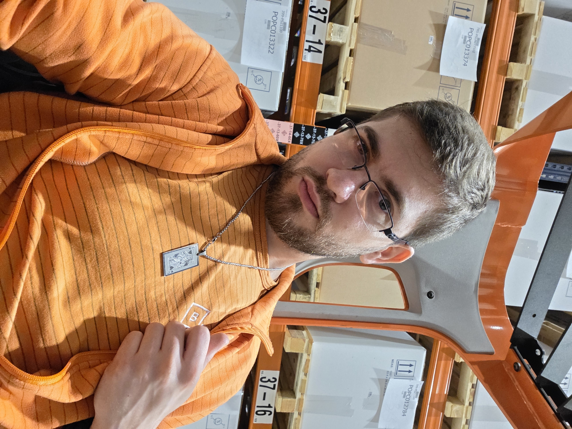 A man with a beard and glasses is standing in a warehouse or storage facility. He is wearing an orange zip-up sweater with a silver necklace. Behind him are shelves of boxes and wooden pallets with labels. The labels show the numbers "37-14," "37-16," and codes like "POPC013374," "POPC013322," and "POPC012784." The location is not explicitly stated as Plovdiv, Bulgaria in the image, but the context of the prompt suggests it might be.