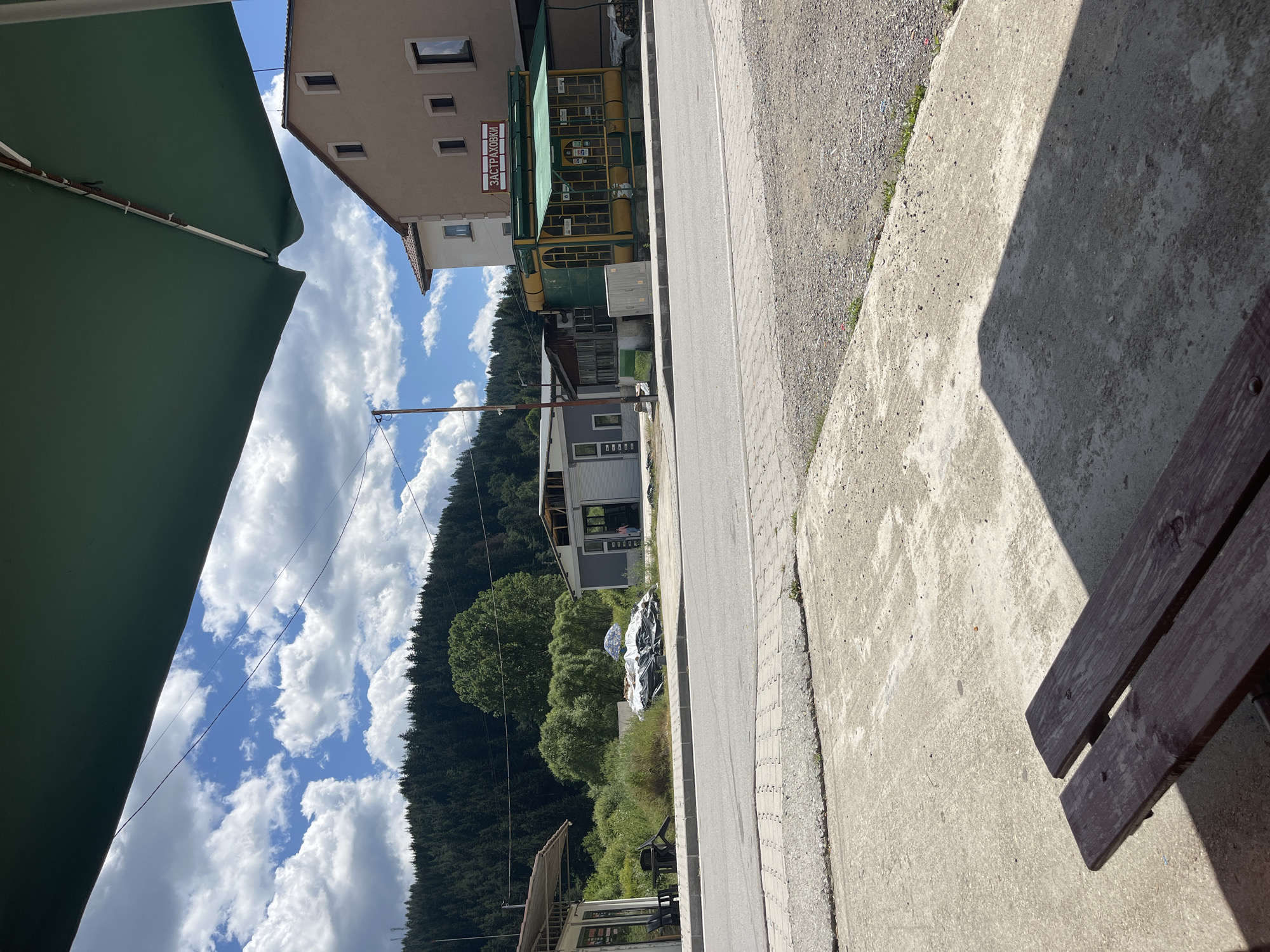 This image depicts a scene in the town of Barutin, Bulgaria, likely near the town center, given the presence of commercial establishments and the main road. In the foreground, a concrete surface and a wooden bench are visible, casting shadows, suggesting an outdoor seating area, possibly for a cafe or rest stop. Beyond this, a paved road runs horizontally across the frame, with a sidewalk or paved area adjacent to it.
On the other side of the road, several buildings and structures are present. To the left, a small, open-fronted kiosk or shop with chairs and tables is partially visible. Next to it, a pile of items covered with tarpaulin and a blue and white umbrella sits on the ground. Further to the right, a gray building with a modern design and what appears to be an entrance is situated.
Dominating the mid-ground is a distinctive yellow and green building with large windows and a green awning. A sign above it reads "ЗАСТРАХОВКИ" (Insurance) in red letters. To the left of this building, a smaller, older structure with wooden elements is attached. Behind these commercial buildings, lush green vegetation and trees form a dense backdrop, leading to a heavily forested hillside that rises towards a bright blue sky dotted with fluffy white clouds. The overall impression is that of a quiet, rural town on a sunny day.