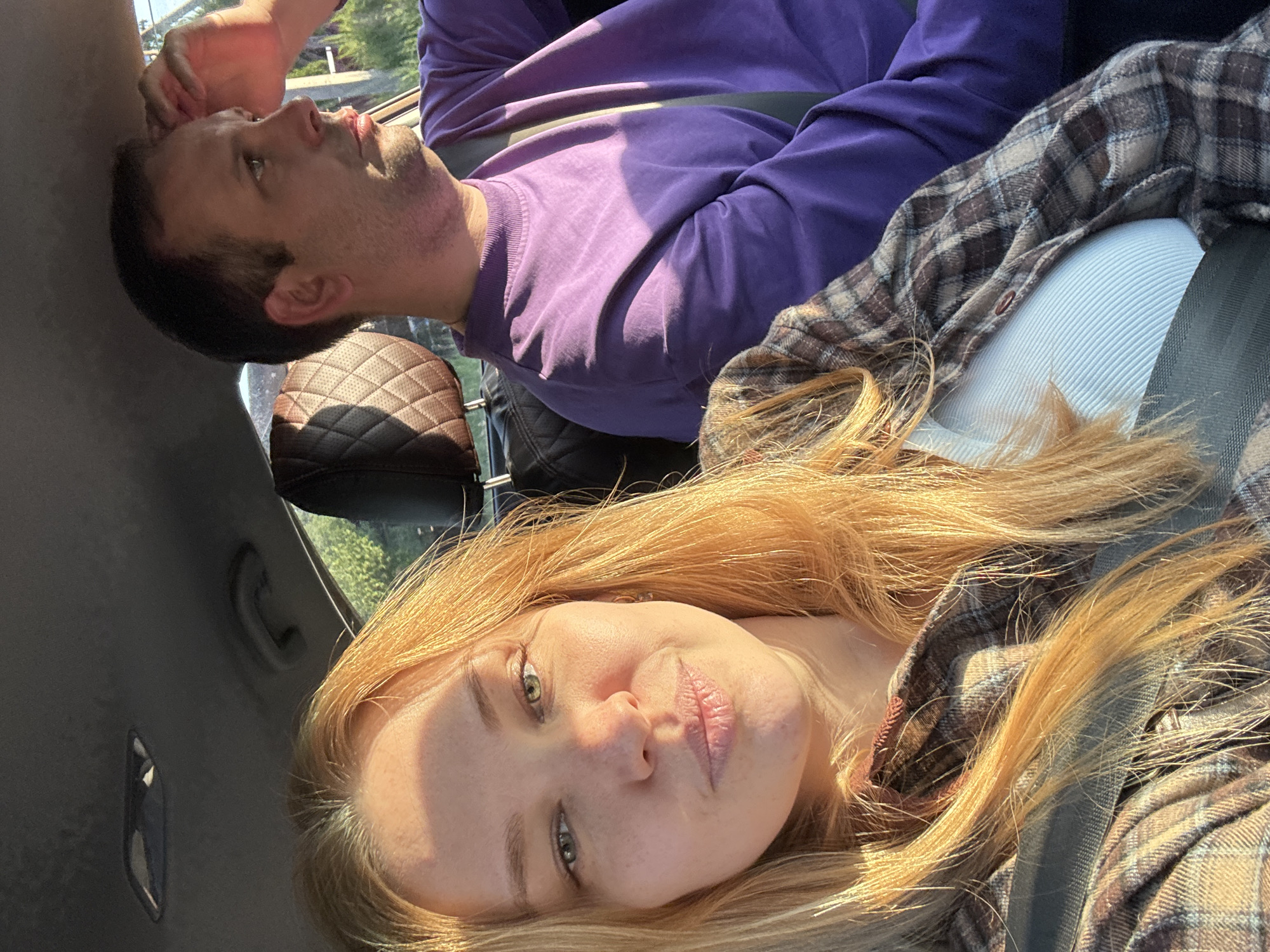 The image features two individuals inside a vehicle, illuminated by bright, warm sunlight. In the foreground, a woman with long, golden-blonde hair and light-colored eyes looks directly towards the viewer with a neutral to slightly pensive expression. Her face is softly lit by a golden glow, highlighting her features. She is wearing a light-colored garment underneath a plaid flannel shirt in shades of brown, grey, and cream, with a seatbelt secured across her.
Behind and above her, a man is visible, looking upwards and to his left, away from the camera. He has short, dark hair and light stubble, wearing a solid purple long-sleeved sweatshirt, also secured by a seatbelt. The interior of the vehicle is evident, including a dark, quilted headrest or seat detail behind the man and parts of the car's roof lining.
The strong sunlight streaming into the vehicle indicates a daytime scene, likely morning or late afternoon, casting distinct shadows and imparting a warm ambiance. The activity suggests a shared journey or outing. The location context for the image is Медягино, Russia. No visible text is present in the image.