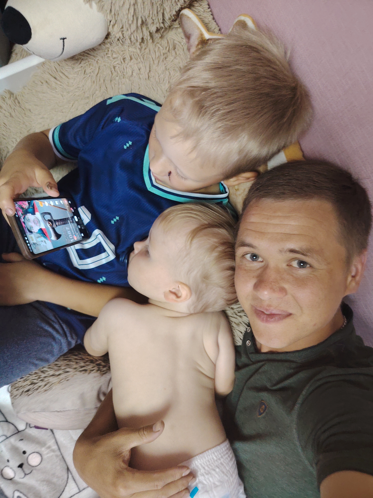 A selfie captures an overhead view of a man, two young children, and stuffed animals on a soft, textured surface that appears to be a bed or couch. The man is on the right side of the frame, looking towards the camera with a slight smile. He has short brown hair and is wearing a dark green collared shirt.
To his left, a baby is lying on its back, facing away from the camera. The baby has blonde hair and is wearing a diaper. The baby's back and head are positioned between the man and the other child.
On the left side of the frame, an older child with blonde hair is looking down at a smartphone held in his hands. He is wearing a blue t-shirt with light blue accents. The phone displays a video feed of a person, possibly a parent, with graphics and text overlays.
Several stuffed animals are visible in the background. A large, light brown shaggy stuffed animal with a black nose and embroidered "J" is at the top left. Another stuffed animal, possibly a fox due to its ears, is partially visible behind the older child's head. A pillow with a hedgehog pattern is beneath the baby. The overall environment is soft and appears to be a domestic setting, likely a child's bedroom or a family living space. The lighting suggests an indoor environment, possibly during the day.
The image is a personal photograph, likely taken by the man himself. There is no information to indicate its location in the city of Chaikovsky, Russia.