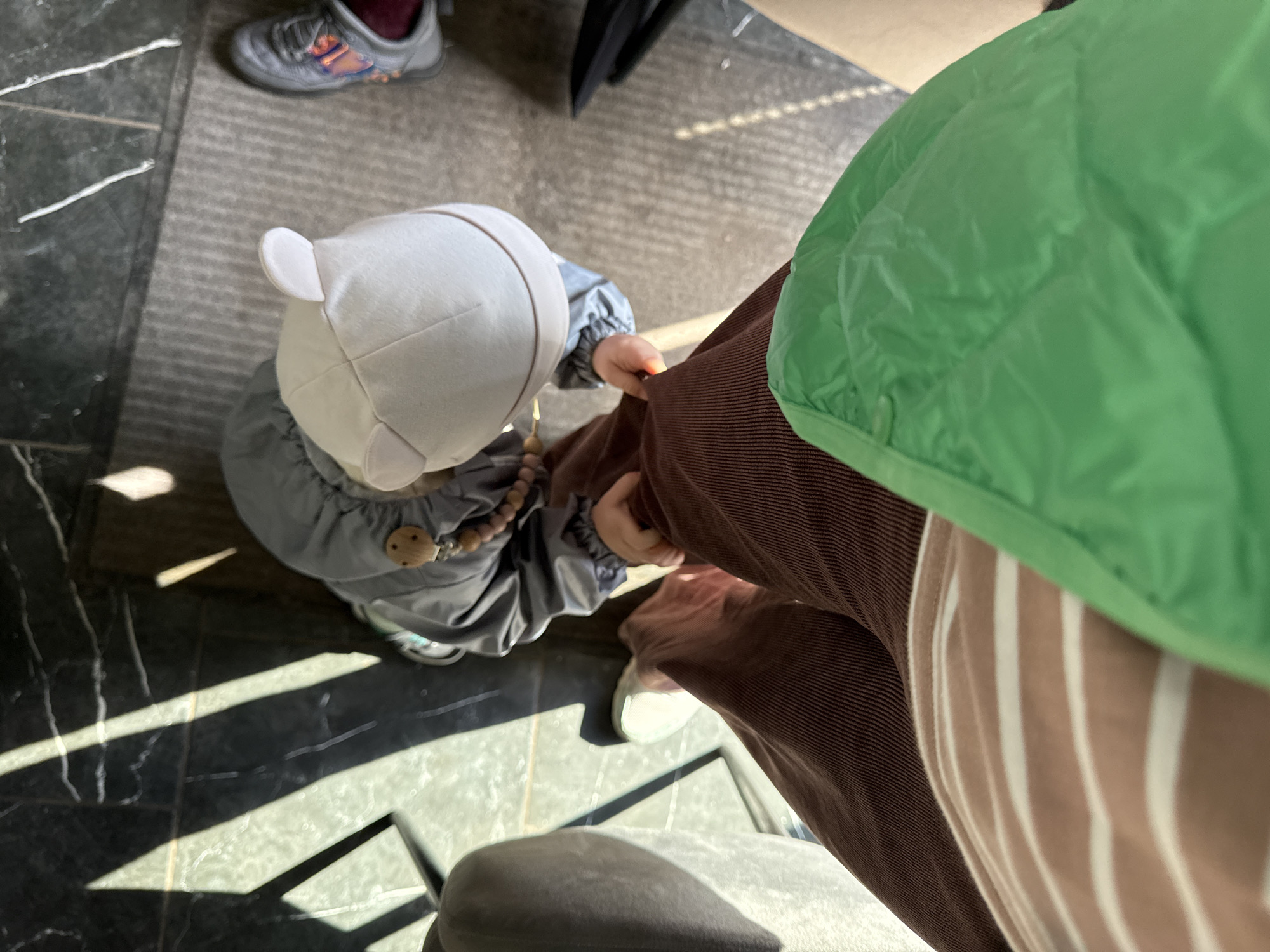 An overhead shot reveals a scene indoors. A baby wearing a white hat and grey outfit is situated to the left. Adjacent to the baby, an individual in brown corduroy pants and a green puffer jacket. Striped fabric is partially visible. To the left, black marble flooring with light stripes. In the background, a grey sneaker. A chair is partially visible. The location is unknown.