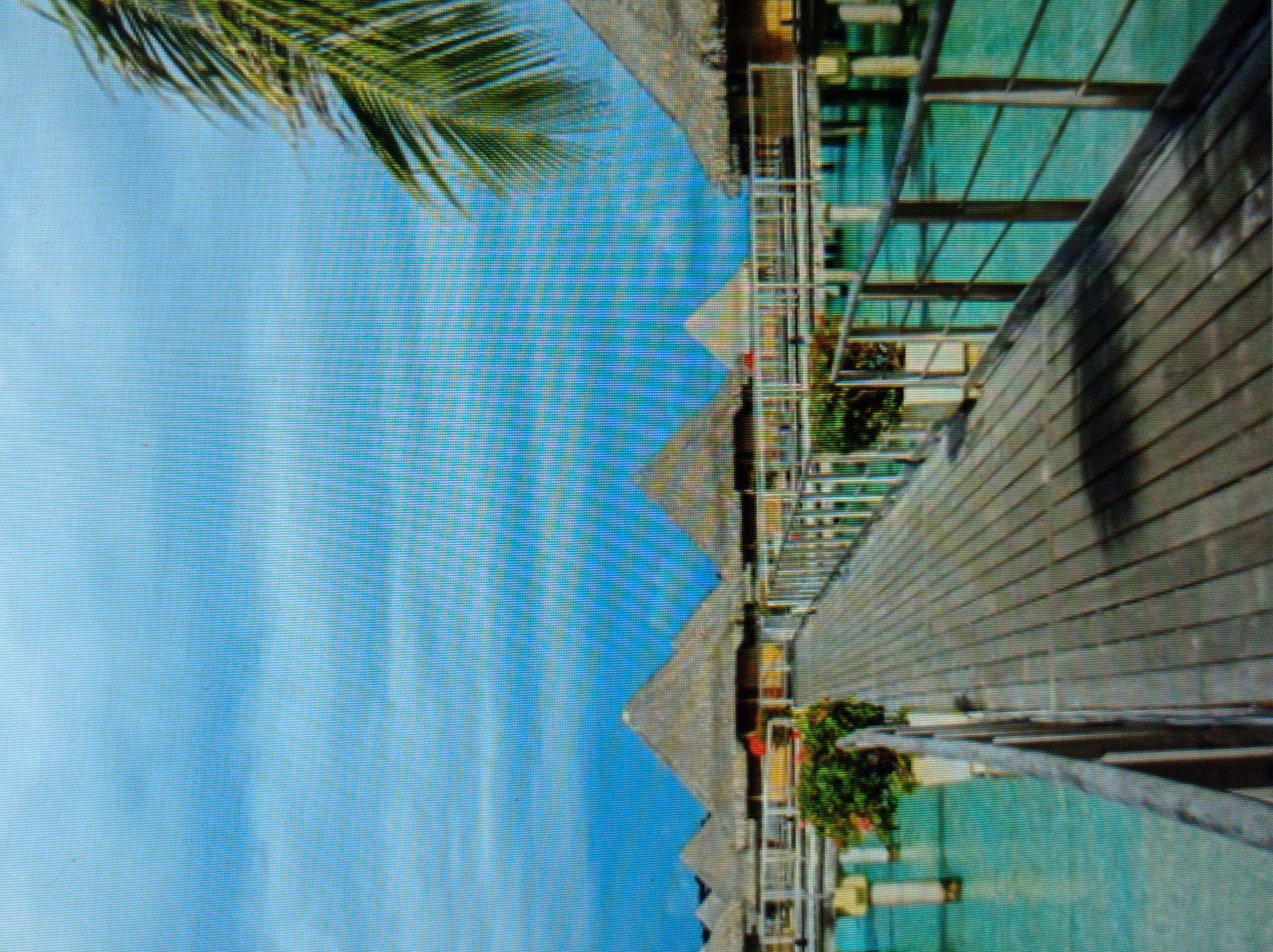 The image displays a serene tropical scene with overwater bungalows and a wooden walkway. The bungalows have thatched roofs and are elevated above clear turquoise water. Lush green foliage and potted plants adorn the walkway, which extends into the distance, leading towards the structures. A palm frond is visible in the upper right corner of the frame, hinting at the tropical environment. The sky is a vibrant blue with scattered clouds. There are no people or animals visible in the image. The scene is peaceful and evokes a sense of a vacation or resort setting. There is no indication or relevance to the city of Mukachevo, Ukraine.