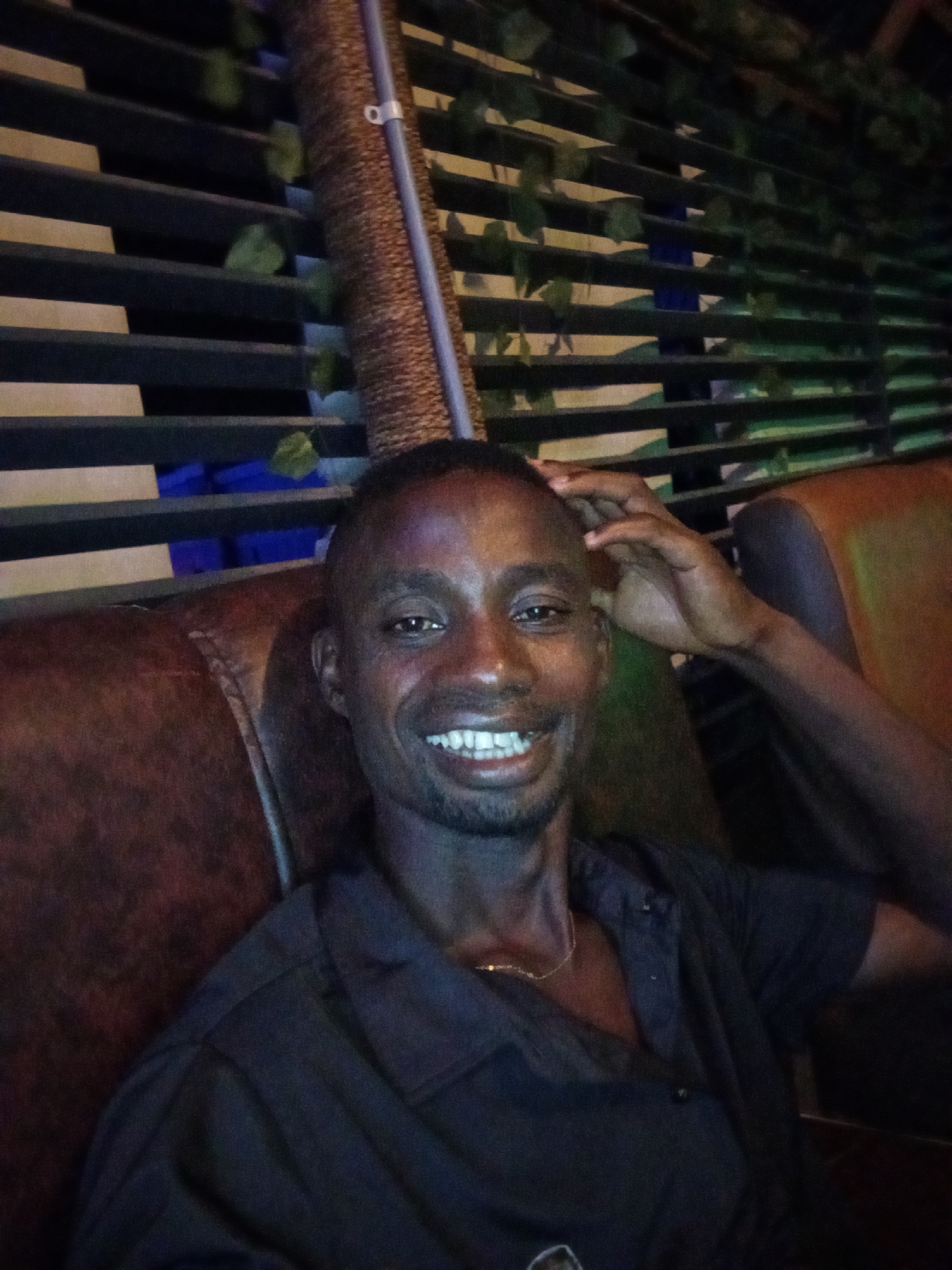 A man is seated, smiling and touching his head. He is wearing a dark button-down shirt and a gold chain is visible around his neck. He is seated on a brown, leather-like surface. Behind him is a decorative wall with horizontal slats and climbing plants. There is a cylindrical structure next to the wall. The scene appears to be indoors. I am unable to verify the location is Adonkolo, Nigeria based on the visual information provided.