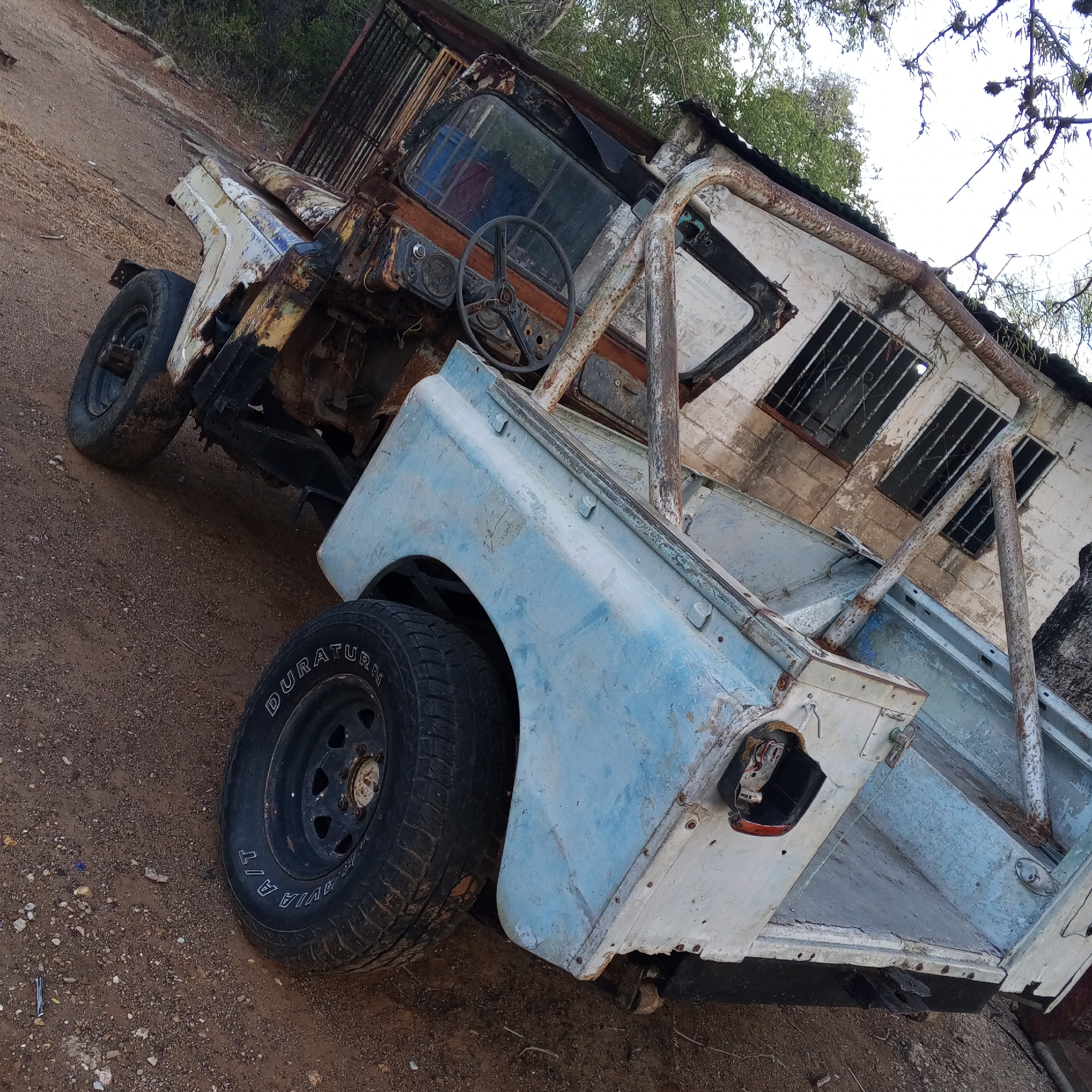 An old, dilapidated vehicle rests on a dirt surface. The vehicle is a jeep or similar off-road type, severely deteriorated with rust and missing parts. The paint is faded and peeling, with a mix of light blue and white. The tires are still present, though worn, with the word "DURATURN" visible on one. The vehicle's interior is heavily damaged, with a visible steering wheel and exposed mechanics. Behind the vehicle is a structure, possibly a building or addition, with barred windows and weathered walls. The surrounding environment consists of dry dirt, sparse vegetation, and some trees in the background. The image suggests a scene of abandonment and decay, likely in a rural or neglected area. While the location is Altagracia, Venezuela, there is no specific city element visible in the provided image.