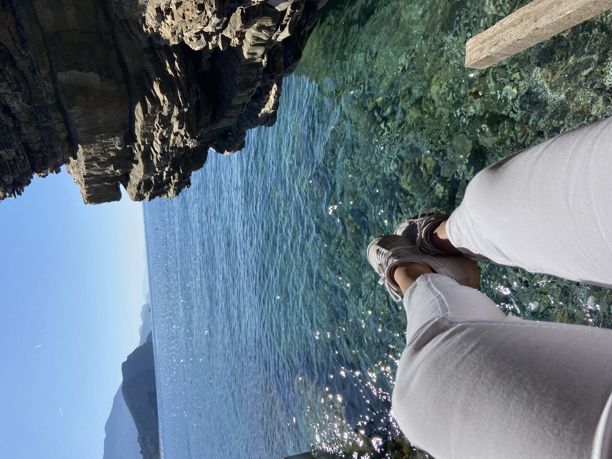 The image depicts a scene at the edge of a clear body of water, likely the Adriatic Sea, near Budva, Montenegro. The viewer's perspective is from a person sitting on a rocky outcrop.
**Subjects:**
* **Human:** The lower portion of a person's body is visible, wearing white pants and neutral-colored sneakers.
* **Environment:** The environment includes the sea, a rocky cliff, and the sky. The water is transparent, revealing the seabed with various stones and pebbles. Mountains are visible in the background.
**Objects:**
* **Clothing:** White pants, neutral-colored sneakers.
* **Natural elements:** Rocks, sea, sky, mountains.
* **Wooden beam:** There is a wooden beam at the upper-right part of the image.
**Actions/Interactions:**
* A person is sitting at the edge of a rock formation overlooking the water.
* The sunlight is interacting with the water, creating sparkling reflections.
**Environment:**
* **Location:** The scene is set in a coastal environment. The presence of mountains in the background indicates a location along the Montenegrin coast, with the crystal-clear water being indicative of the Adriatic Sea.
* **Weather/Lighting:** The scene is well-lit, suggesting a sunny day.
**City of Budva Context:**
The scene is likely located near Budva due to the presence of clear waters, rocky coastlines, and mountainous landscapes characteristic of this region in Montenegro. This area is known for its scenic beauty and rocky shores, which are consistent with the image's depiction.