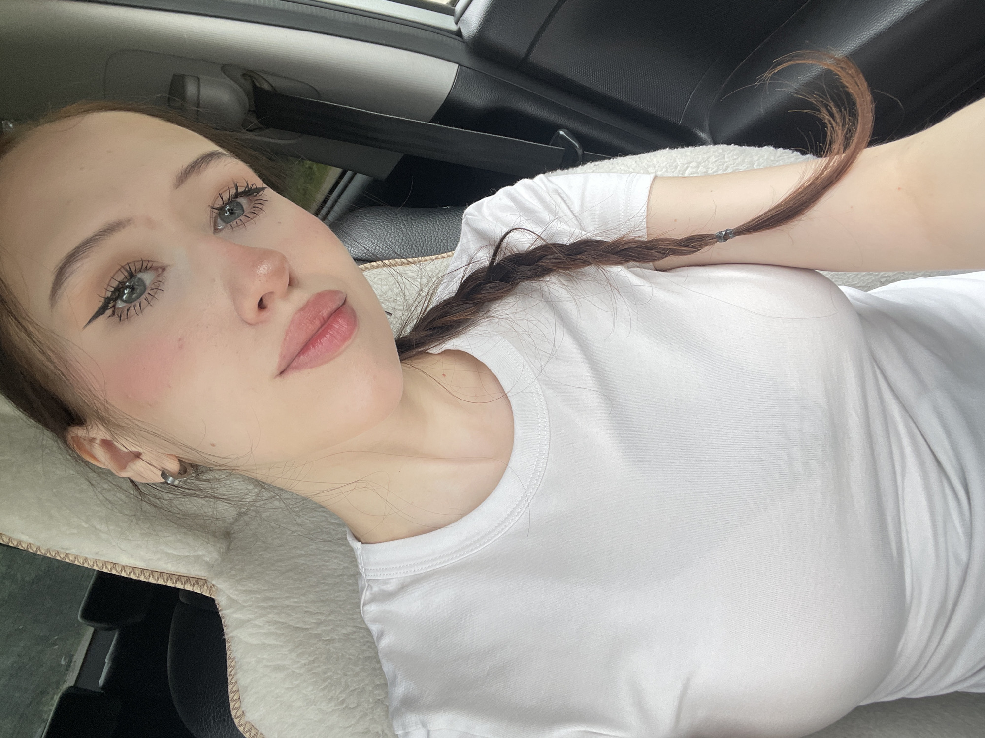 In Gavrilovka, Russia, a young woman with fair skin, light blue eyes, and dark brown hair braided into a pigtail sits in the passenger seat of a vehicle. She is wearing a plain white t-shirt. Her makeup includes winged eyeliner, mascara, and blush. The car's interior is visible, with a fuzzy white seat cover behind her and a black seat and seatbelt to her right. The focus is on her face and upper body as she takes a selfie.