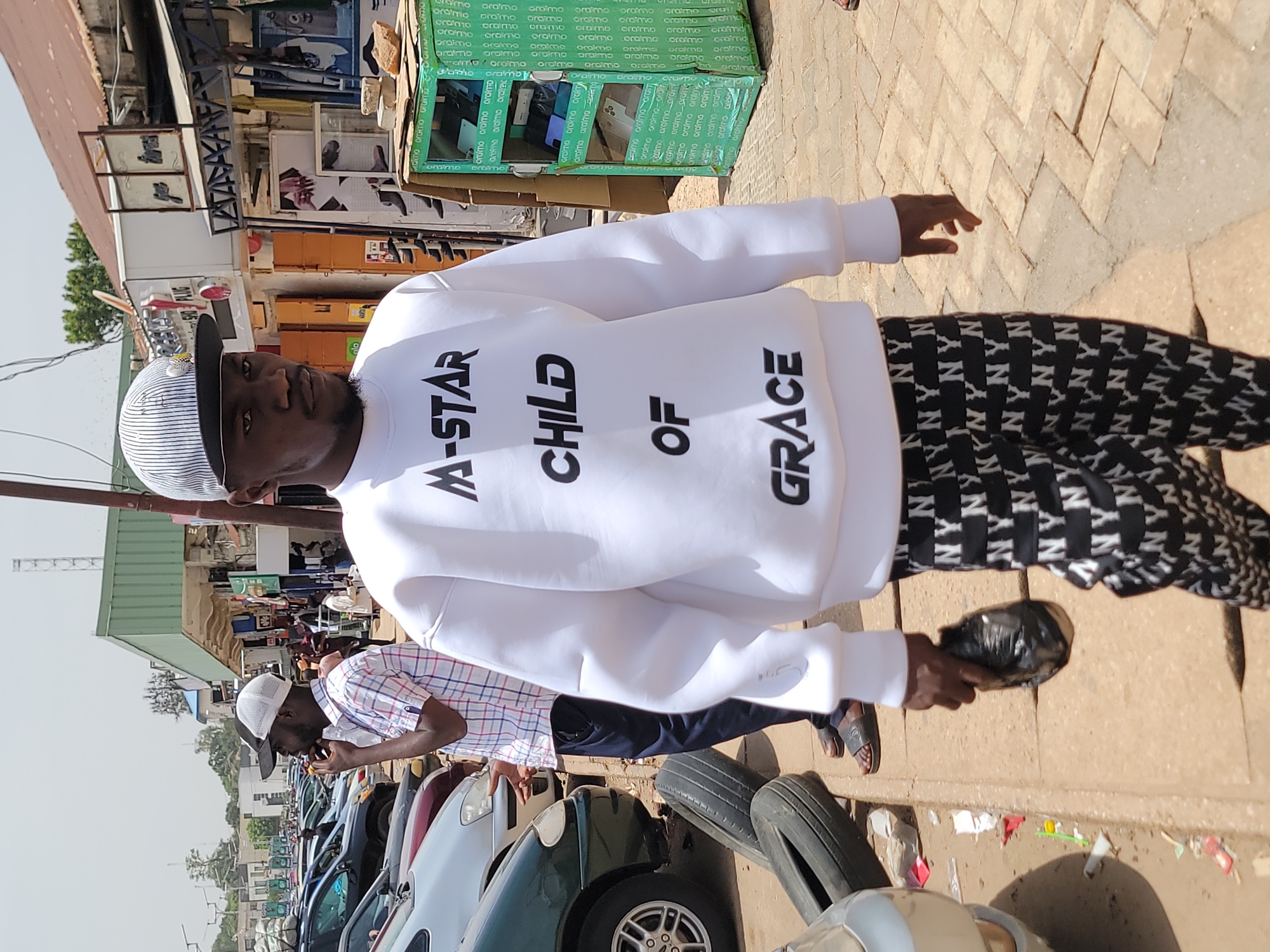 A man stands on a city sidewalk wearing a white sweater printed with "M-STAR CHILD OF GRACE" in black letters and black and white patterned pants. He is holding a black plastic bag. In the background, there is a row of parked cars, another man eating from a brown paper bag, and a store displaying phones behind glass with the word "oraimo" repeated across the glass. Boxes of phones are piled nearby. The sidewalk is paved with interlocking blocks. The location is likely Jos, Nigeria.