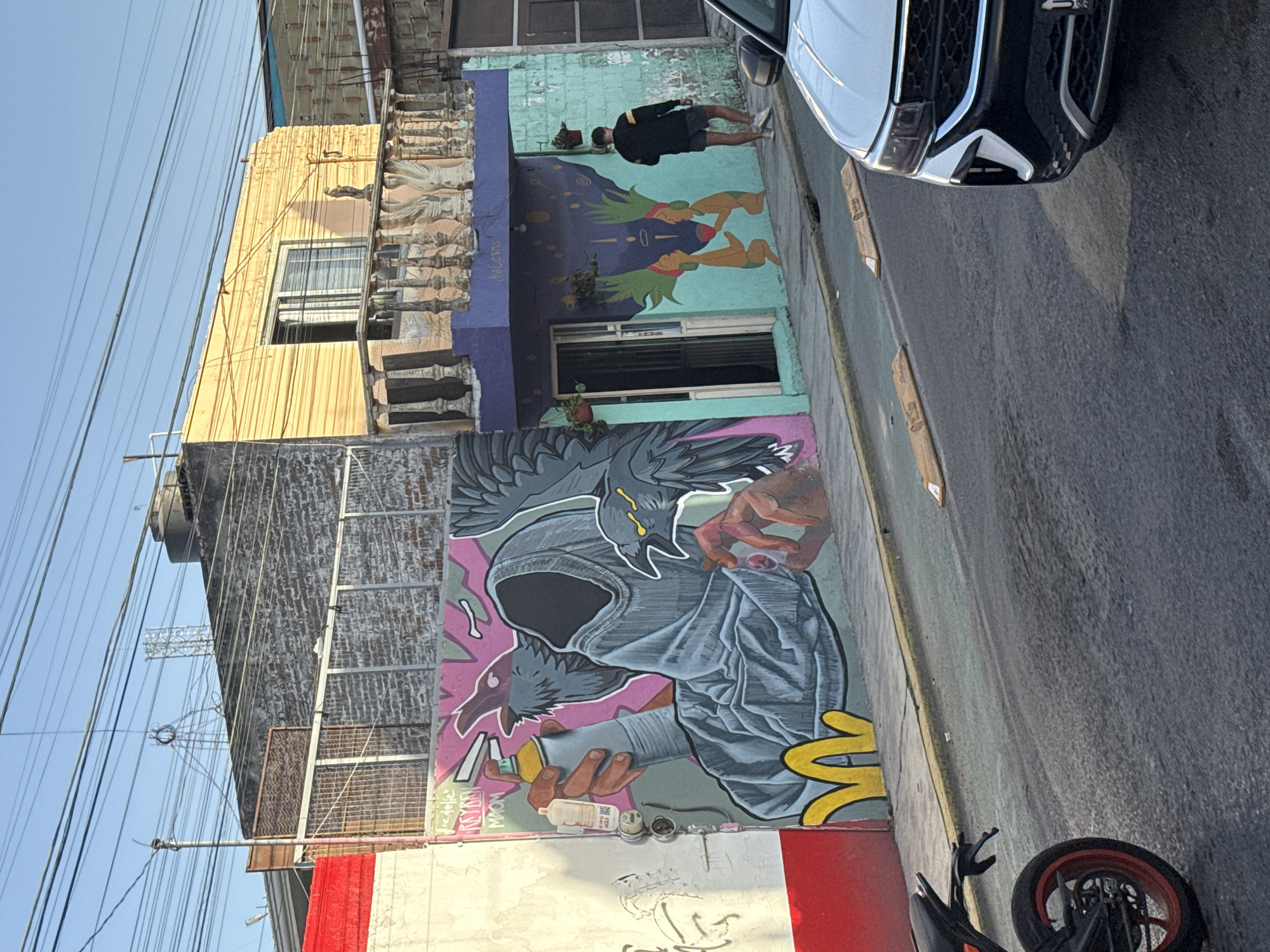 The image captures a street scene in San Andrés Cholula, Mexico, prominently featuring urban art. A multi-story building with various architectural styles, including a yellow section with linear detailing and a more ornate section with decorative moldings, forms the backdrop. Murals adorn the building's walls. One mural depicts figures with stylized features and vibrant colors, while another portrays a hooded character with avian elements, holding a spray can.
A white vehicle with black accents occupies the upper right portion of the frame, partially obstructing the view. The street is visible, showcasing a motorcycle parked near the building. Overhead, power lines extend across the sky. A person is standing on the sidewalk in front of the building.