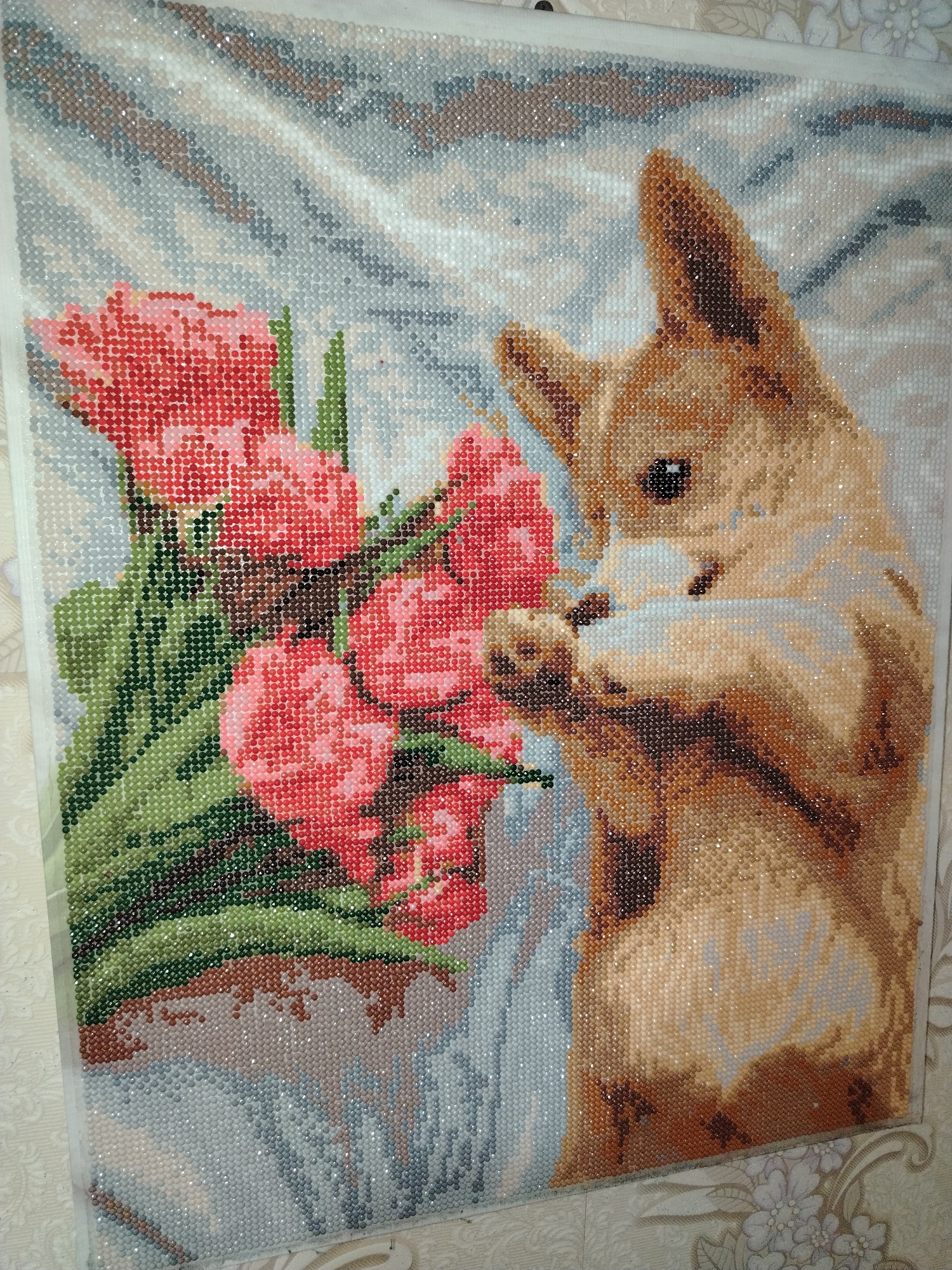A rectangular diamond painting is depicted, showing a stylized scene of a light brown and tan dog with a bouquet of red flowers. The dog, positioned on the right, faces left, its front paws raised towards the flowers as if sniffing them. Its fur is rendered in various shades of brown, tan, and cream diamond beads, featuring a dark eye and nose. The bouquet, on the left, consists of multiple full-petaled red or deep pink flowers with green leaves and stems, arranged in a light-colored vase or container. The background of the artwork is a soft, diffused pattern of light blue, grey, white, and subtle brown diamond beads. The diamond painting is resting against or mounted on a wall covered with light-colored wallpaper featuring a repeating floral and vine pattern in cream, beige, and light grey tones. Unfinished canvas edges of the diamond painting are visible, particularly along the top and left. The location is Unknown, Unknown.