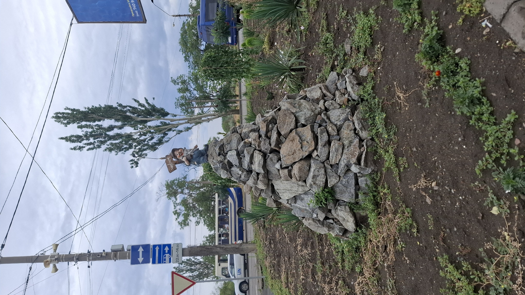 A statue of a cartoon character holding a "welcome" sign stands atop a pile of rocks. The statue is a masculine figure with brown hair, wearing a brown hat, blue shirt, and blue overalls. The pile of rocks is located in a landscaped area with green plants and dark soil. In the background, a white car with "UN" markings is parked on the left, and a blue bus is driving past. A utility pole with multiple speakers and signs is visible on the left. The sky is cloudy. The scene appears to be outdoors in an urban or suburban environment.