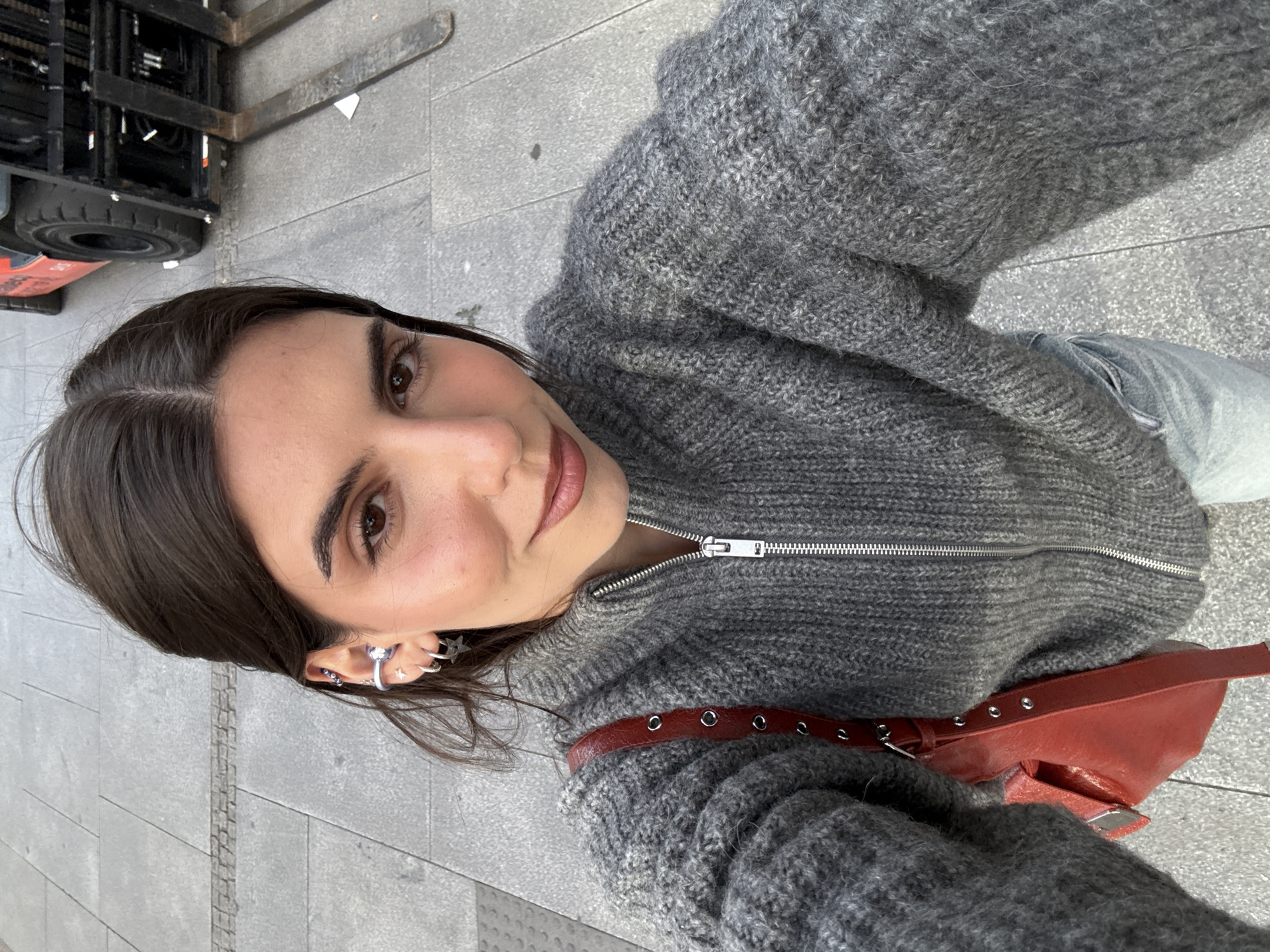 A selfie depicts a woman with brown hair and eyes, wearing a gray zip-up sweater and light-wash jeans. She has multiple silver earrings in her left ear. A red leather bag with a belt-like strap rests on her right side. The background reveals a gray concrete sidewalk, a portion of a forklift truck with black tires and orange accents, and a blurred building facade, suggesting an urban environment. Although the exact location cannot be determined with certainty from the provided information, the combination of urban features could potentially align with an industrial or commercial zone in Madrid, Spain, if that is indeed the actual location.