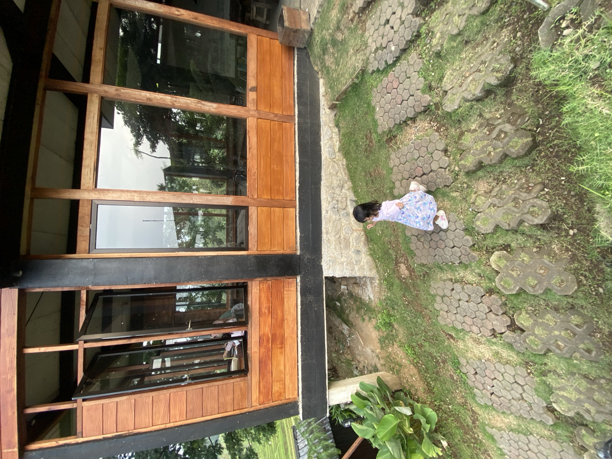 The image shows a young child in a light-colored dress with a floral pattern walking along a paved pathway next to a building. The building has a wooden facade with large windows. The pathway is made of gray hexagonal pavers and flower-shaped stones, with grass growing in between the stones. The location is in Tamiajeng, Indonesia.