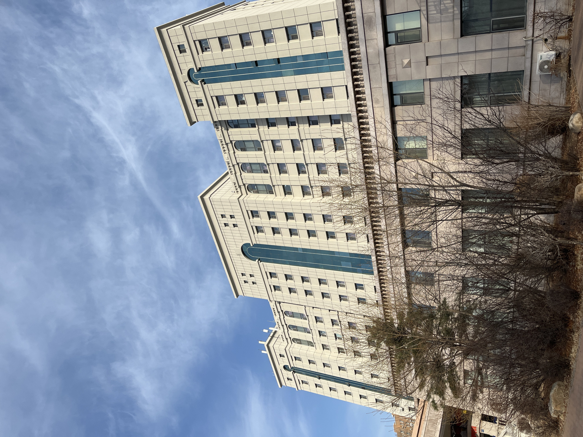 The image shows two tall buildings in Changchun City, China. One building is a light gray color with a teal blue stripe down the center. The other building is a darker gray color. There are bare trees and bushes in front of the buildings. The sky is blue with wispy white clouds. The perspective is from a low angle, looking up at the buildings.