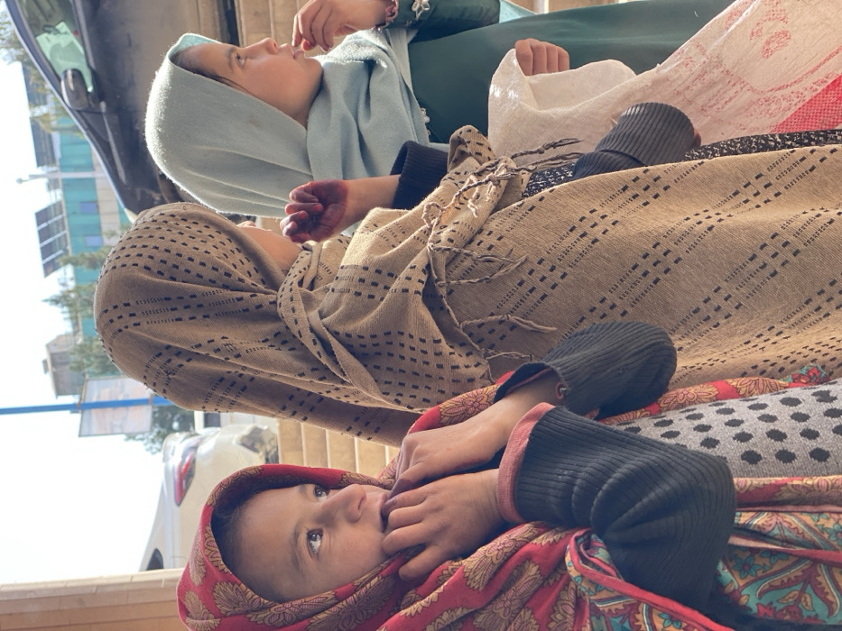 Three young girls are depicted standing in a street scene in Kandahar, Afghanistan, under bright daylight. The girl on the left, viewed in a three-quarter profile facing slightly right and upwards, wears a red headscarf with gold and teal patterns, a dark ribbed long-sleeved shirt, and a grey polka-dotted top; her hands are positioned near her mouth. The central girl, primarily viewed from the side and back, wears a light brown shawl with dark rectangular patterns and fringes; her visible left hand displays dark reddish staining, indicative of henna, on the palm and fingers. The girl on the right, seen in profile, wears a light blue headscarf and a dark green long-sleeved dress; her right hand is near her mouth, and she holds a piece of white fabric in her left hand. The background features a street with a partial view of a white car on the left and a dark car on the right, urban buildings with visible rooftop solar panels, and a blue pole structure under a clear sky.