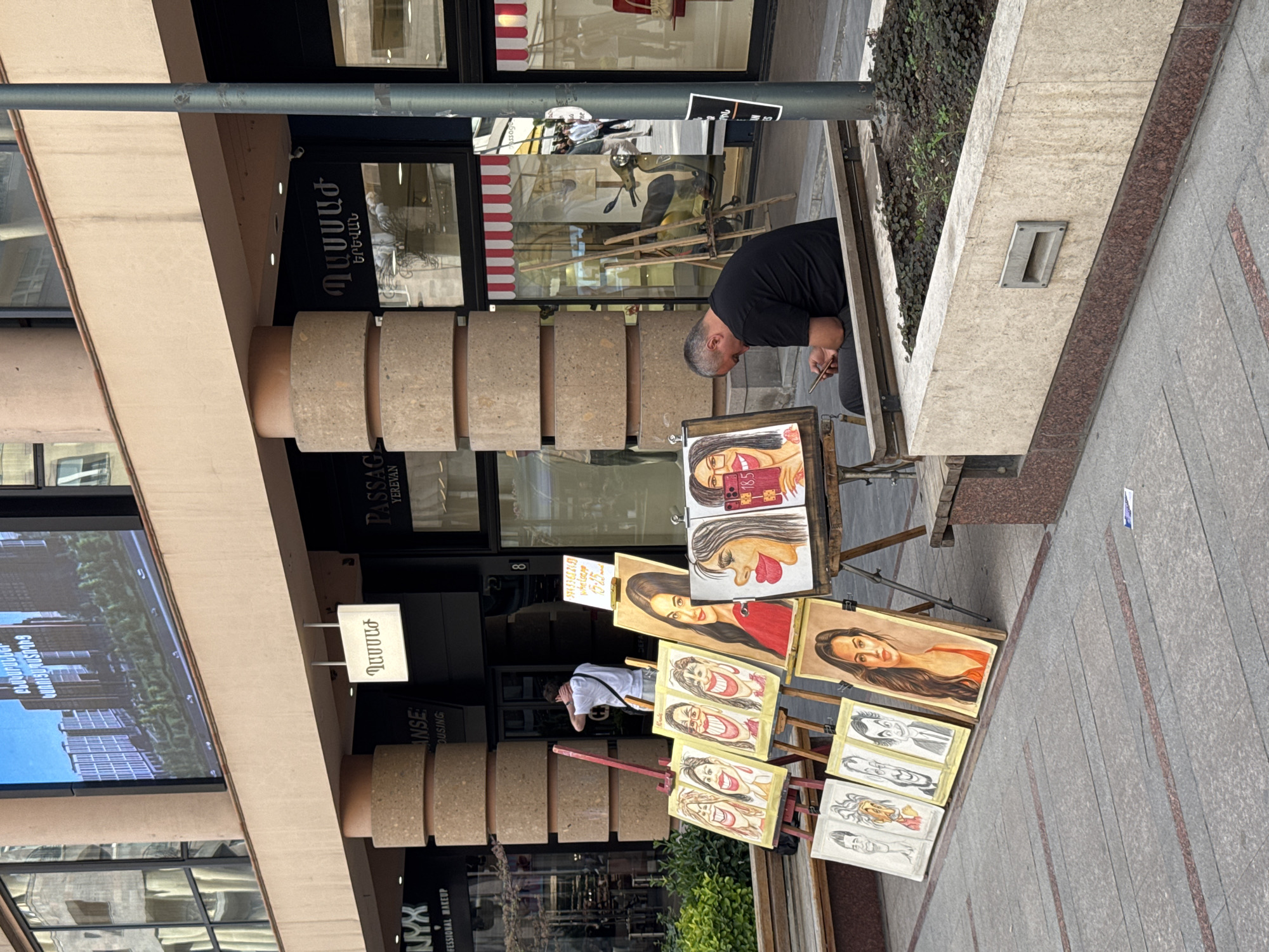 The image depicts an outdoor scene in Yerevan, Armenia. A man is working on artwork displayed on an easel, likely creating or selling portraits. The artwork includes caricatures and portraits of individuals, including one with the number "185" and another holding a phone. The man is focused on his work, and there is a sidewalk with grey and reddish-brown tiles.
The environment includes storefronts behind the artwork, one with the Armenian word "ԲՆԱԿԱՐԱՆՆԵՐ" and the phrase "ԿԱՌՈՒՑԱՊԱՏՈՂԻՑ," which translates to "Apartments from the builder." There are signs indicating "PASSAGE Yerevan" and "NYX Professional Makeup." The number "8" can be seen on one of the storefronts. A planter box with greenery is located near the artist. A passerby is seen looking through the paintings, possibly considering a purchase.