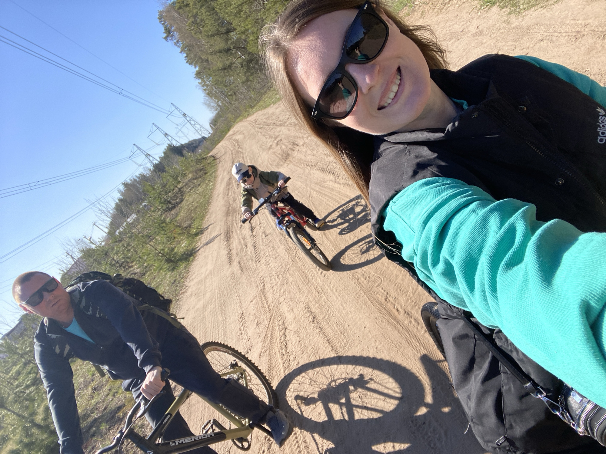 The image shows a selfie taken on a sandy road in Noviy, Russia. Three people are present: a woman taking the selfie, a man to her left, and a child further ahead on the road. All three are riding bicycles. The woman is wearing sunglasses, a black vest, and a turquoise top. The man is also wearing sunglasses and a dark jacket with a backpack. The child is wearing a cap and light-colored jacket. Power lines and towers are visible on the left side of the image, suggesting an area outside of the city center. The weather appears sunny and clear. Tire tracks are visible in the sandy road.