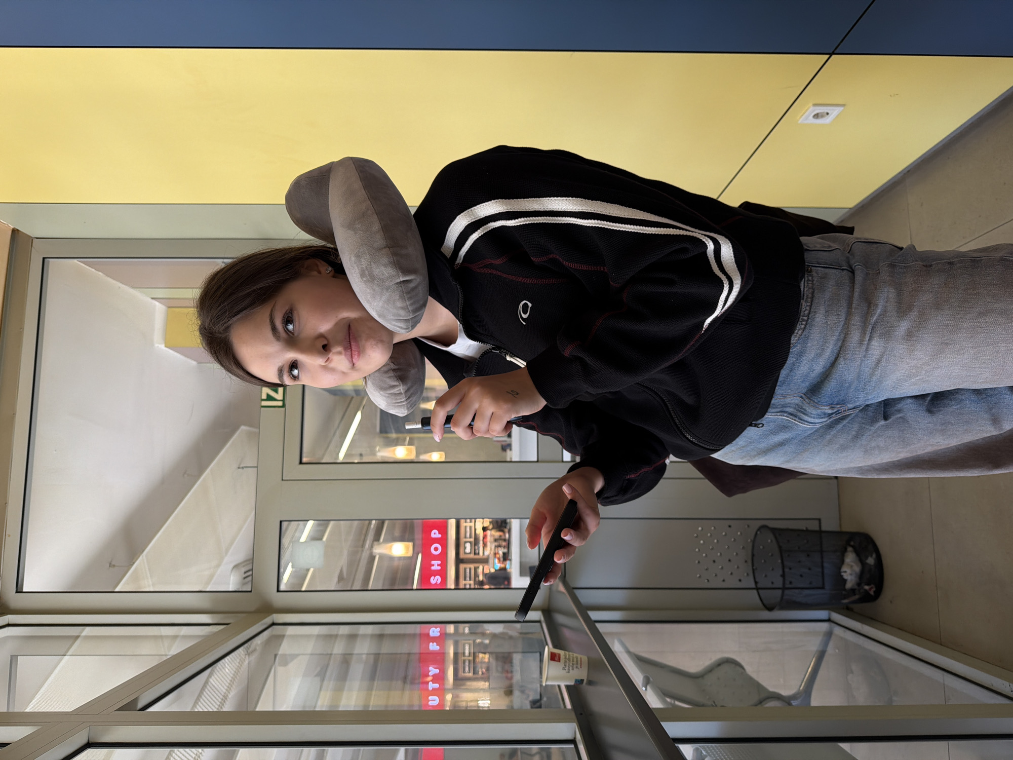 A young woman stands indoors, wearing a gray neck pillow, a black zip-up jacket with white stripes, and light blue jeans. She holds two electronic devices in her hands. Behind her is a glass wall with a reflection of a "BEAUTY FR SHOP" sign. Through the glass, an interior retail space is visible. To the right, there's a mesh wastebasket and a chair. The environment has yellow and blue walls. The scene is likely at an airport, but I lack the information to specify the location in Gornje Dubrave, Bosnia and Herzegovina.