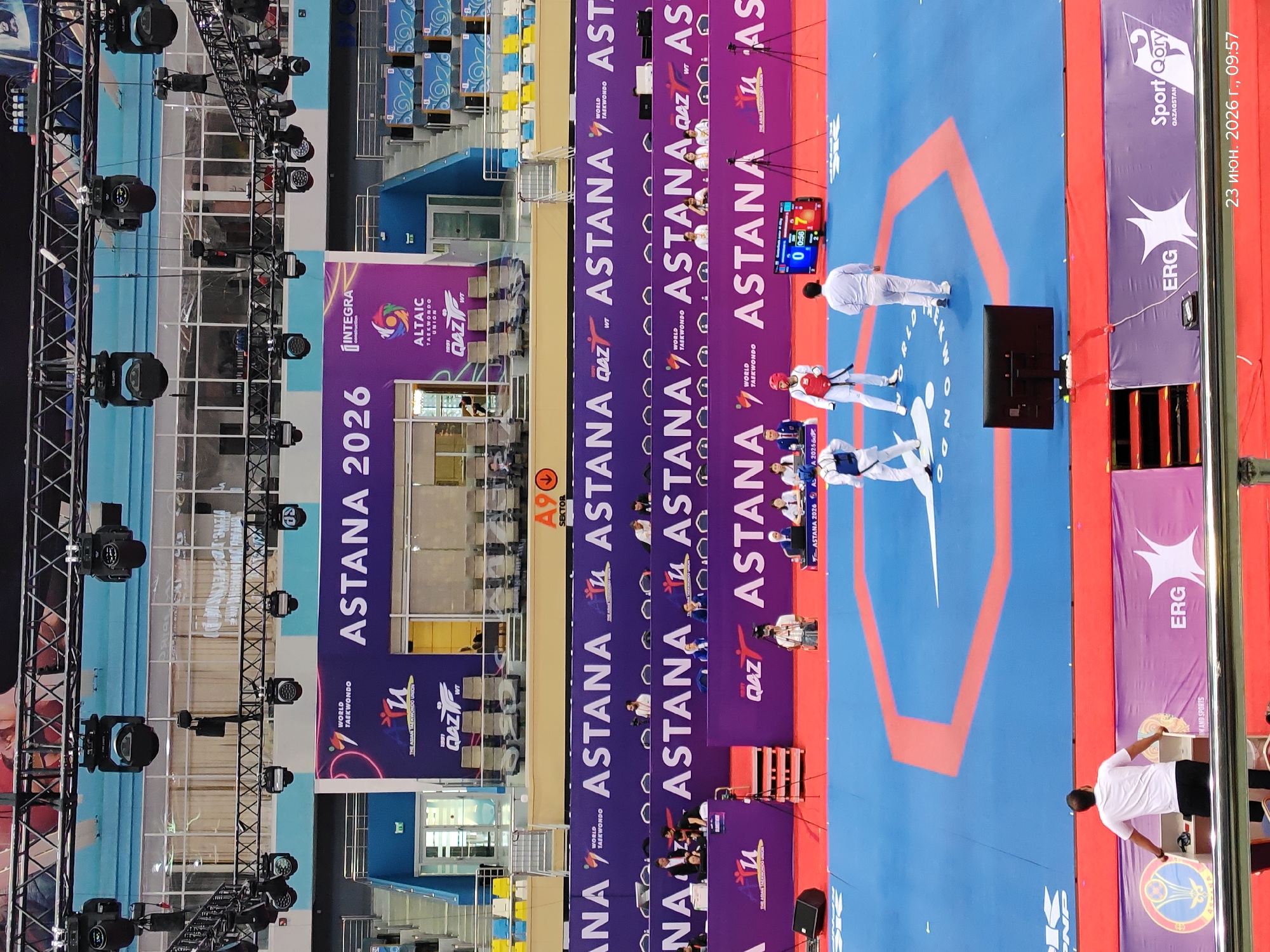 The image captures an indoor taekwondo competition in Astana, Kazakhstan, specifically at the Astana 2026 event. In the foreground, two taekwondo athletes are engaged in a match on a blue and red mat. One athlete, wearing a red and white uniform and a red helmet, is positioned facing left, while another athlete, in a white uniform and white helmet, is facing right. A referee, dressed in white, stands near the center of the mat. A large electronic scoreboard displays the score as "0 0:58 7".
In the mid-ground, spectators are seated in tiered blue and yellow seating areas. Several officials are present behind a purple barrier, some wearing white shirts and ties. A camera operator with a camera on a tripod is also visible.
The background features banners with the "ASTANA 2026 WORLD TAEKWONDO" logo and other sponsor branding, indicating the location and event. The overall environment is a modern sports arena with bright overhead lighting and a clean, well-maintained floor. The date stamp in the bottom right corner reads "23 июня. 2026 г., 09:57", confirming the event is taking place on June 23, 2026.