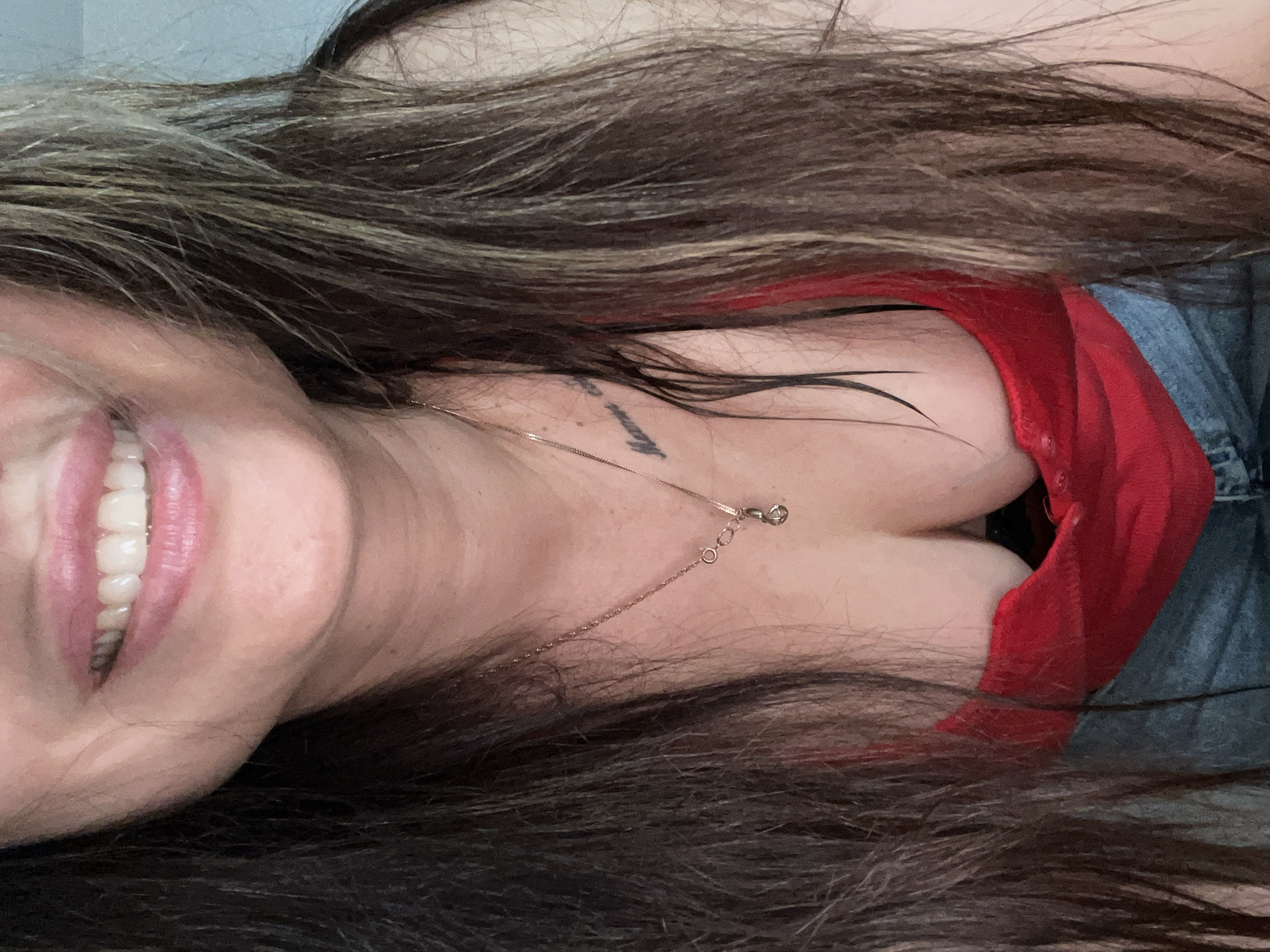 The image, originally oriented sideways, depicts a close-up of a smiling woman, seen from an upward angle. Her long, dark brown hair, interspersed with lighter highlights, frames her face and flows across her shoulders. She wears a bright red top with a deep V-neck or scoop neck, which reveals cleavage. A thin, gold-toned necklace with a small, simple pendant adorns her neck and chest. On her upper chest, partially obscured by hair and the necklace, is a small, dark tattoo, appearing to be text that is not fully legible, possibly starting with 'Never'. A broad smile on her face reveals her upper teeth, conveying an emotion of happiness or joy. To the right, a section of blue denim fabric is visible, possibly part of jeans or a jacket. The background is a plain, light-colored surface, indicative of an indoor environment. The lighting is soft, and the overall impression is one of cheerfulness.