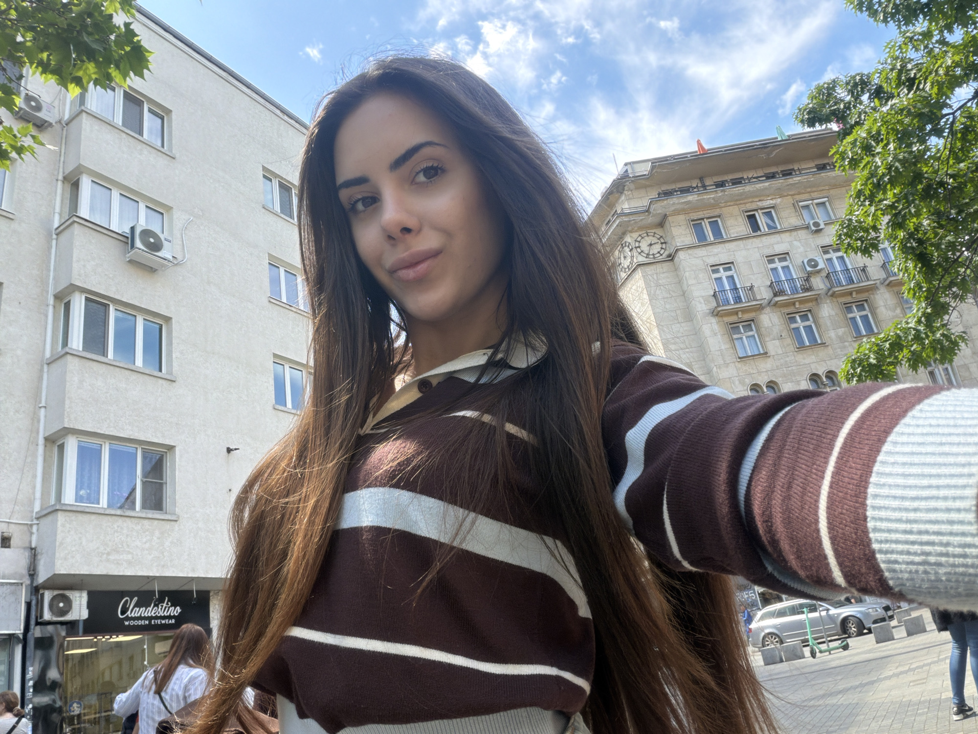 A young woman takes a selfie on a sunny day in Sofia, Bulgaria. The woman has long, dark brown hair and is wearing a brown and white striped sweater. Behind her, there is a building with an air conditioning unit on the side and a store called "Clandestino Wooden Eyewear" on the ground floor. Further behind her, on the right, there is a light-colored building with a clock. People and parked cars are visible on the sidewalk in the background.