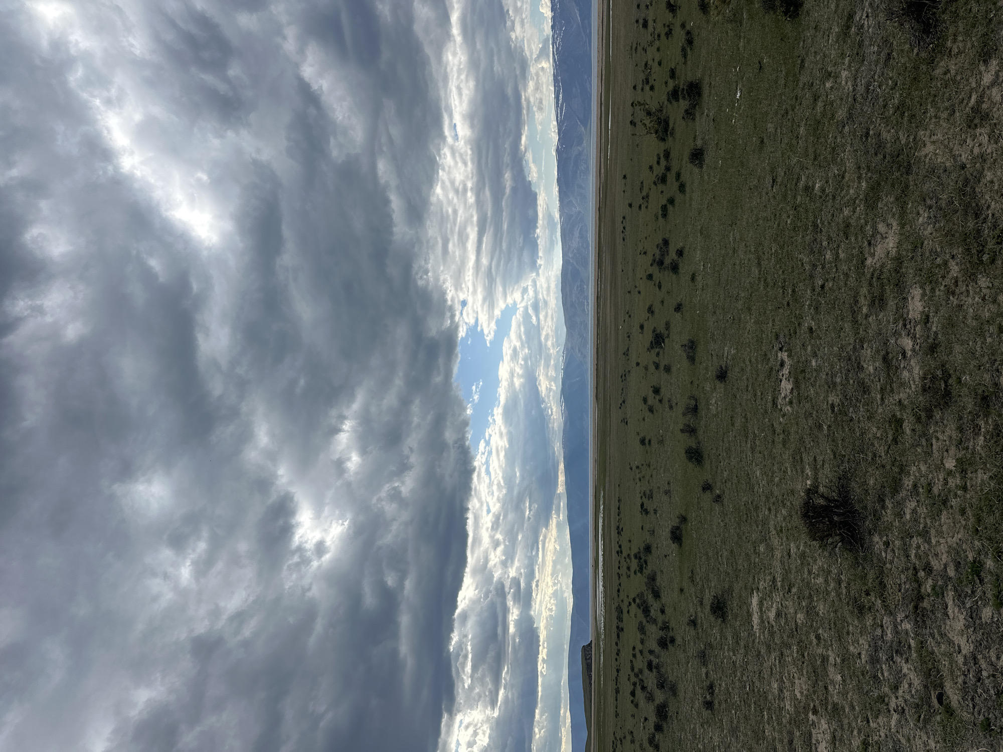 The image depicts a landscape scene. The sky is filled with dark, ominous clouds on the left side, contrasting with a brighter, sunlit area in the center, and lighter clouds on the right. A mountain range is visible in the middle distance, appearing as a narrow strip between the sky and the foreground. The foreground consists of a field, primarily green with patches of brown, indicating possibly dry or dormant vegetation. No people, recognizable subjects, actions, or interactions are discernible in the image. The location, Khuzhir, Russia, is not directly confirmed by the image content but is indicated as a known detail.