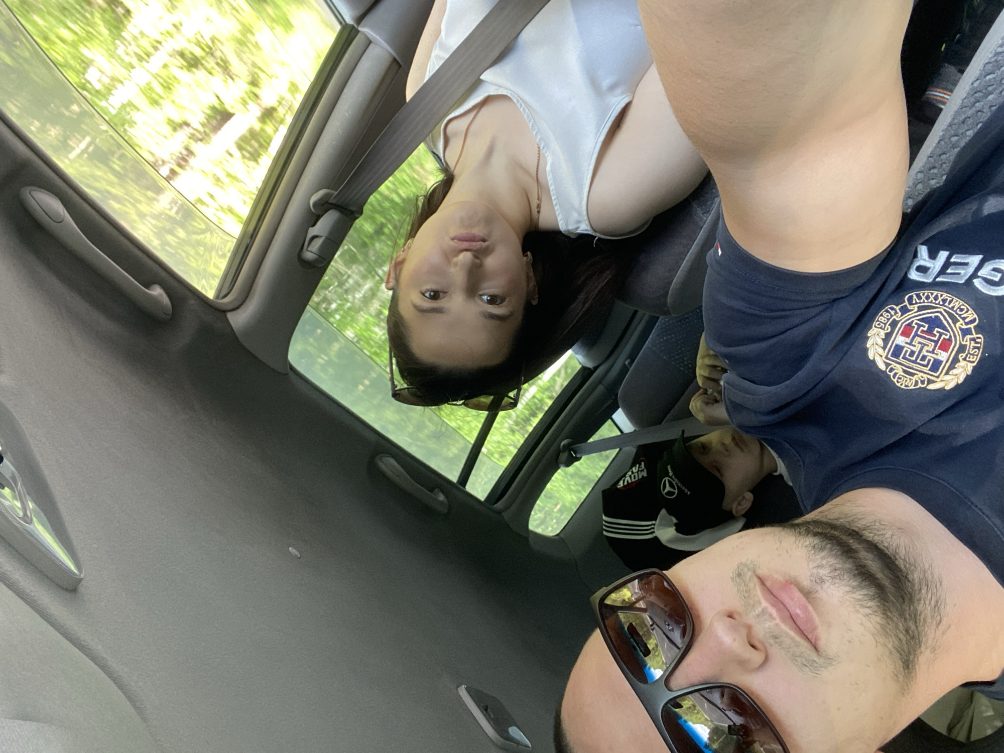A selfie is taken from the perspective of the driver of a vehicle traveling through a wooded area. In the foreground, the driver, a man wearing sunglasses and a dark t-shirt with a logo, is partially visible. To his right, in the back seat, a woman wearing a white top makes a playful facial expression. A young boy, wearing a black baseball cap with a Mercedes-Benz logo, is also seated in the back. The car has a sunroof open, revealing the green trees and foliage blurred by motion outside. The interior of the car appears to be a typical passenger vehicle. The location is identified as Burdakovka, Russia.