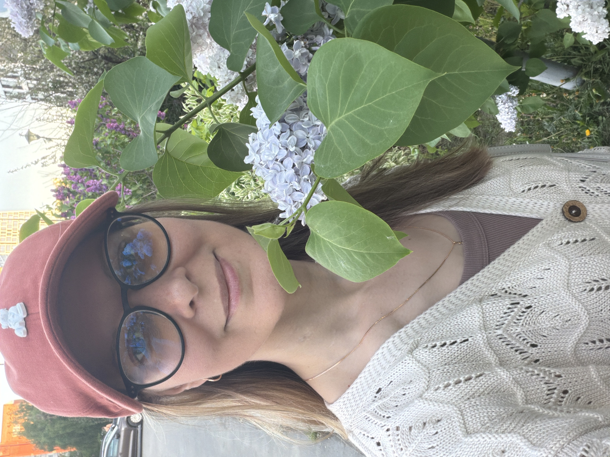 A person is pictured in an outdoor setting with flowering plants. The person is wearing a pink cap with a small white decoration, black-rimmed glasses with reflections in the lenses, a beige top, and a light-colored knit cardigan with a button closure. They are wearing a gold necklace. They are surrounded by green leaves and lilac flowers of varying shades of white and light purple. The surroundings suggest an urban environment, possibly Izhevsk, Russia. In the background, out of focus, are glimpses of buildings and what appears to be a parked vehicle.